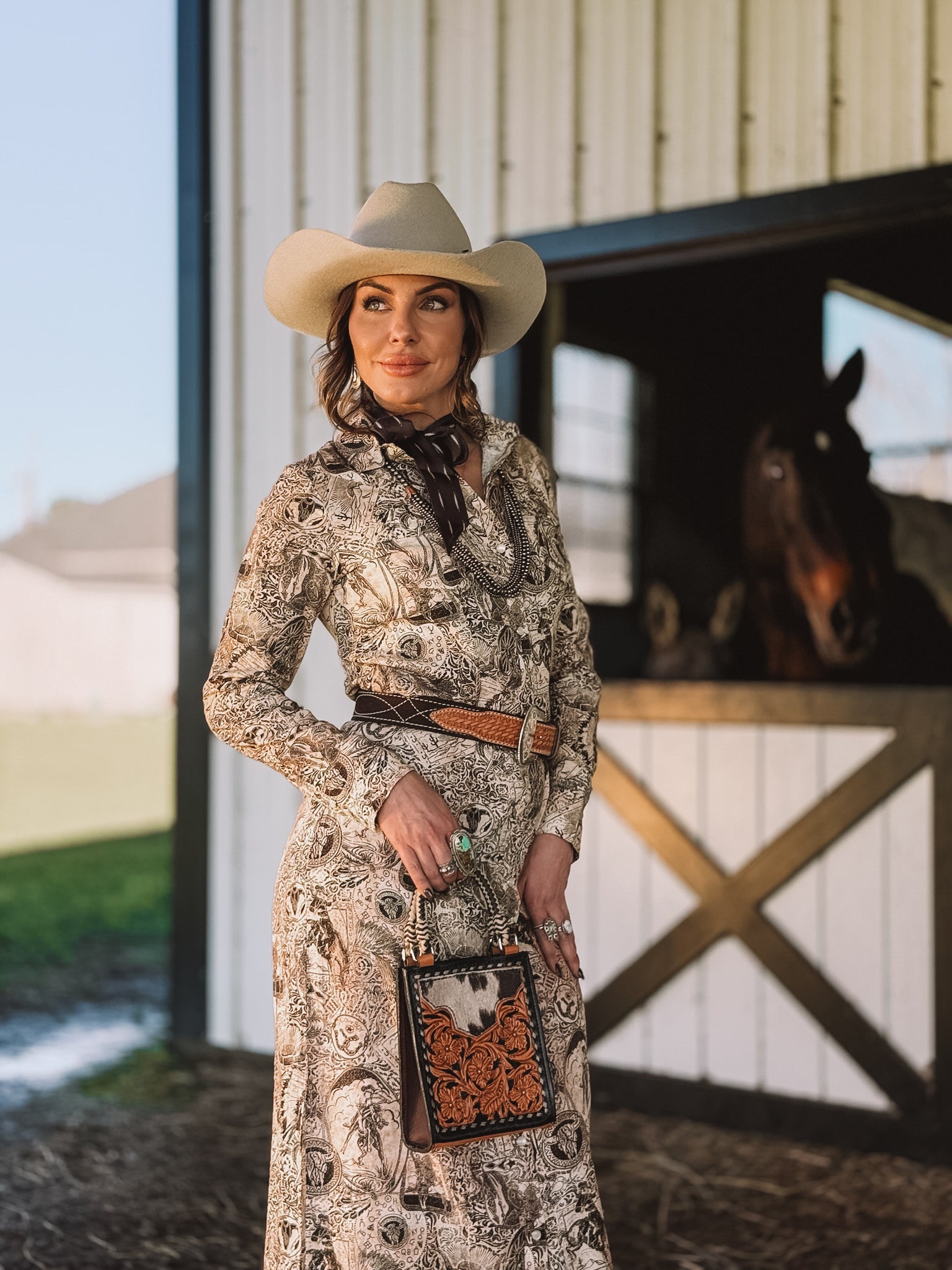 Woman in western boho dress with cowboy hat and tooled leather purse by horse stable