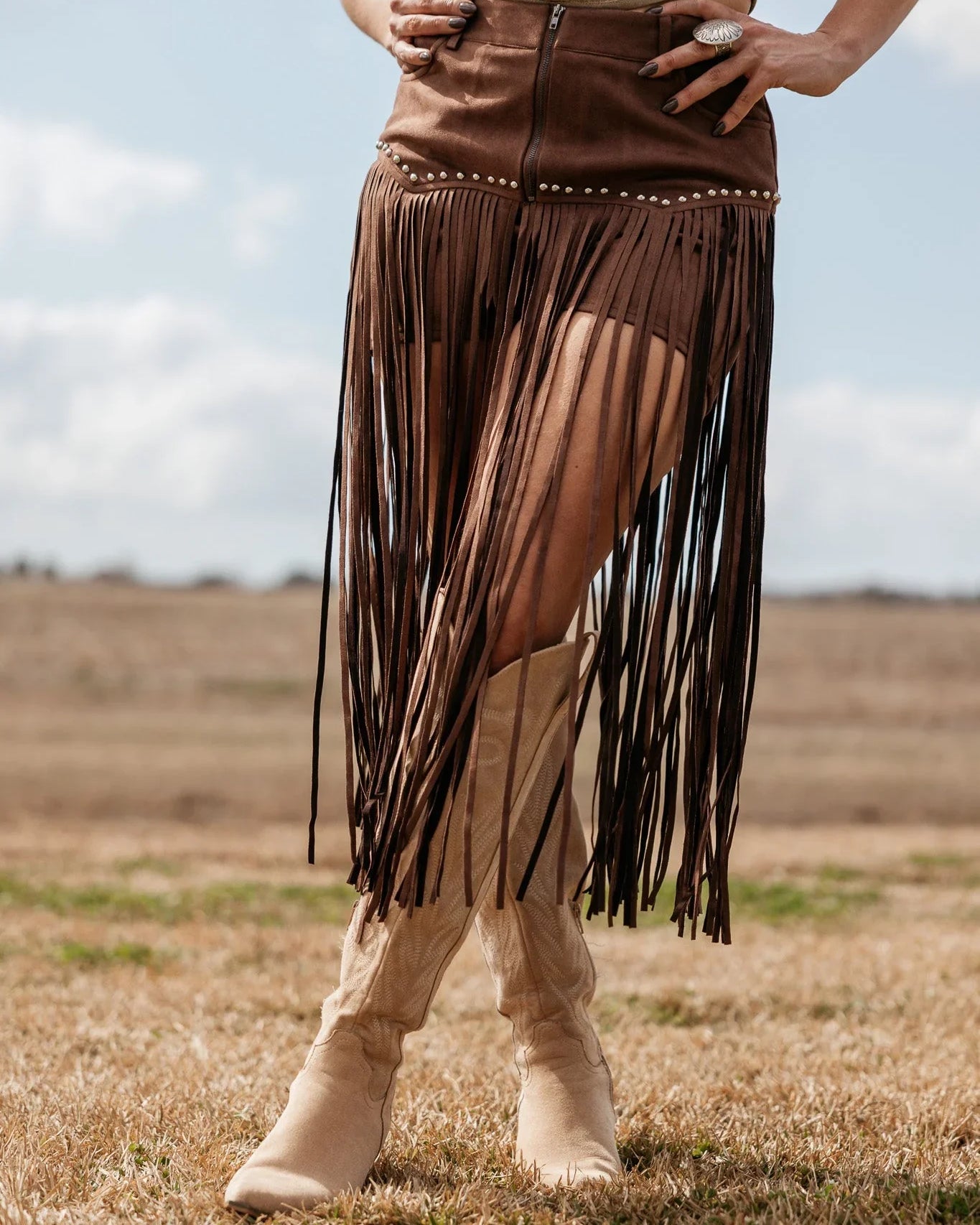 Woman wearing brown fringe suede skirt, western graphic top, and tan cowgirl boots outdoors