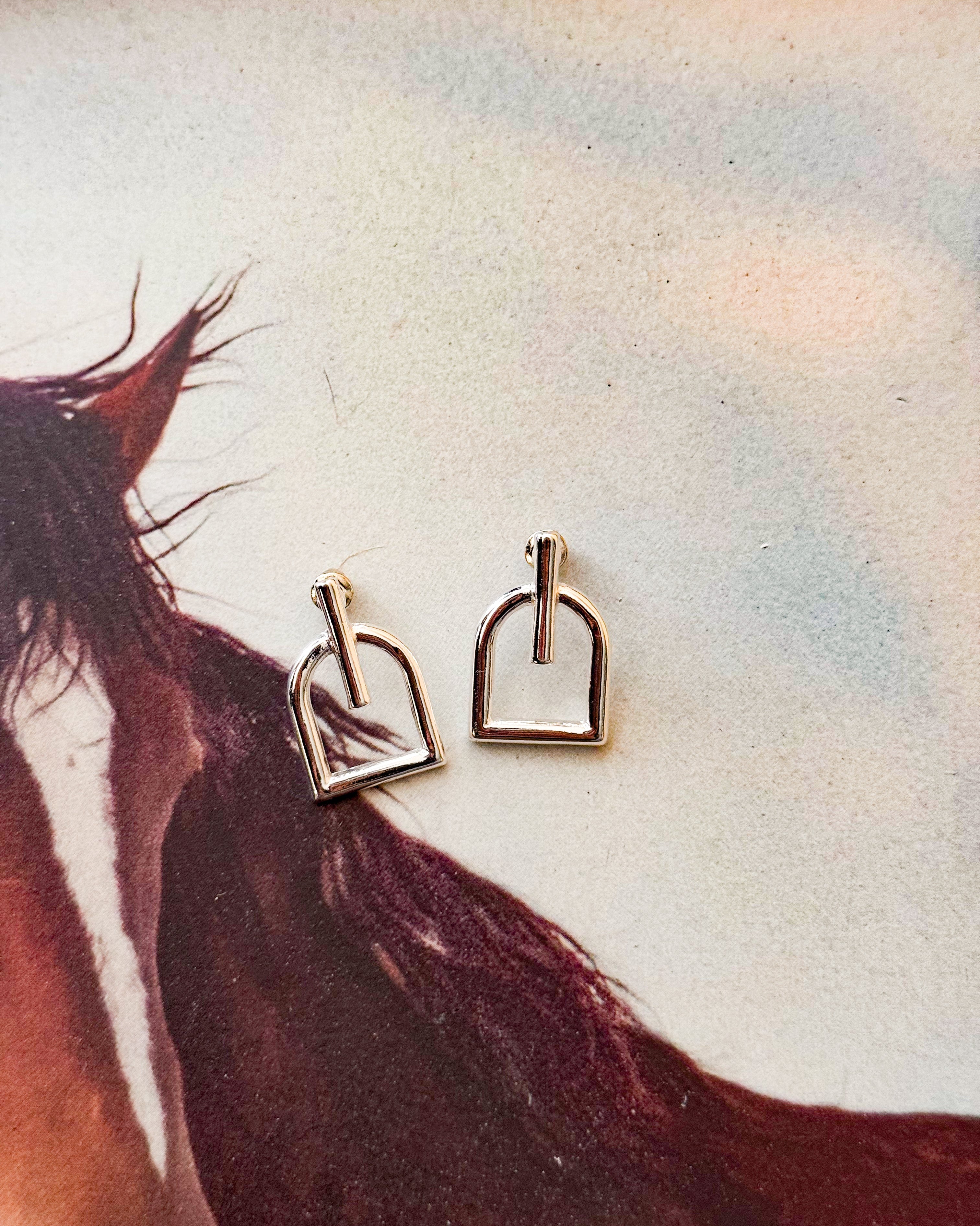 Silver western stirrup earrings on a horse-themed background, boho cowgirl jewelry.