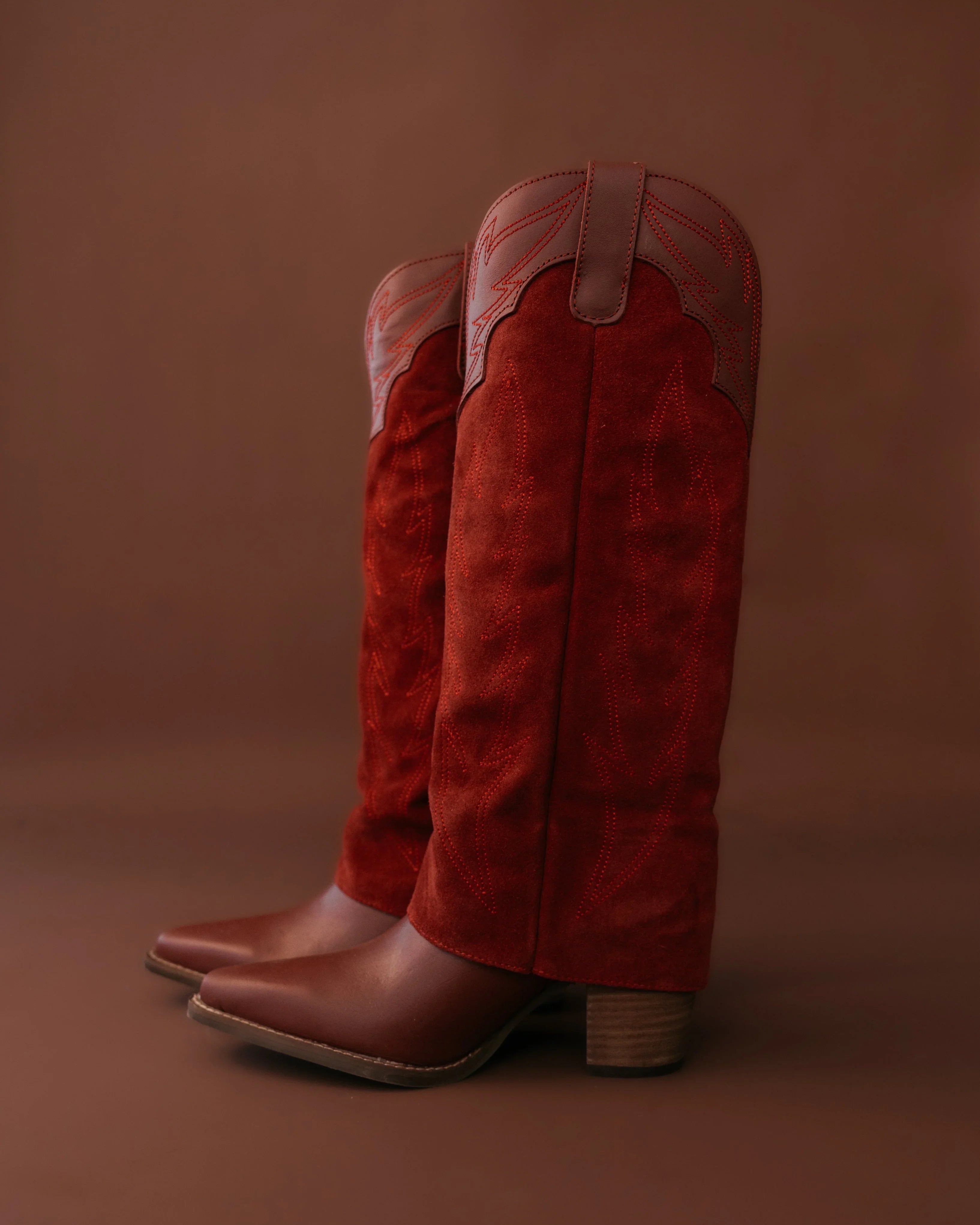 Red suede western cowgirl boots with decorative stitching and block heel on brown background