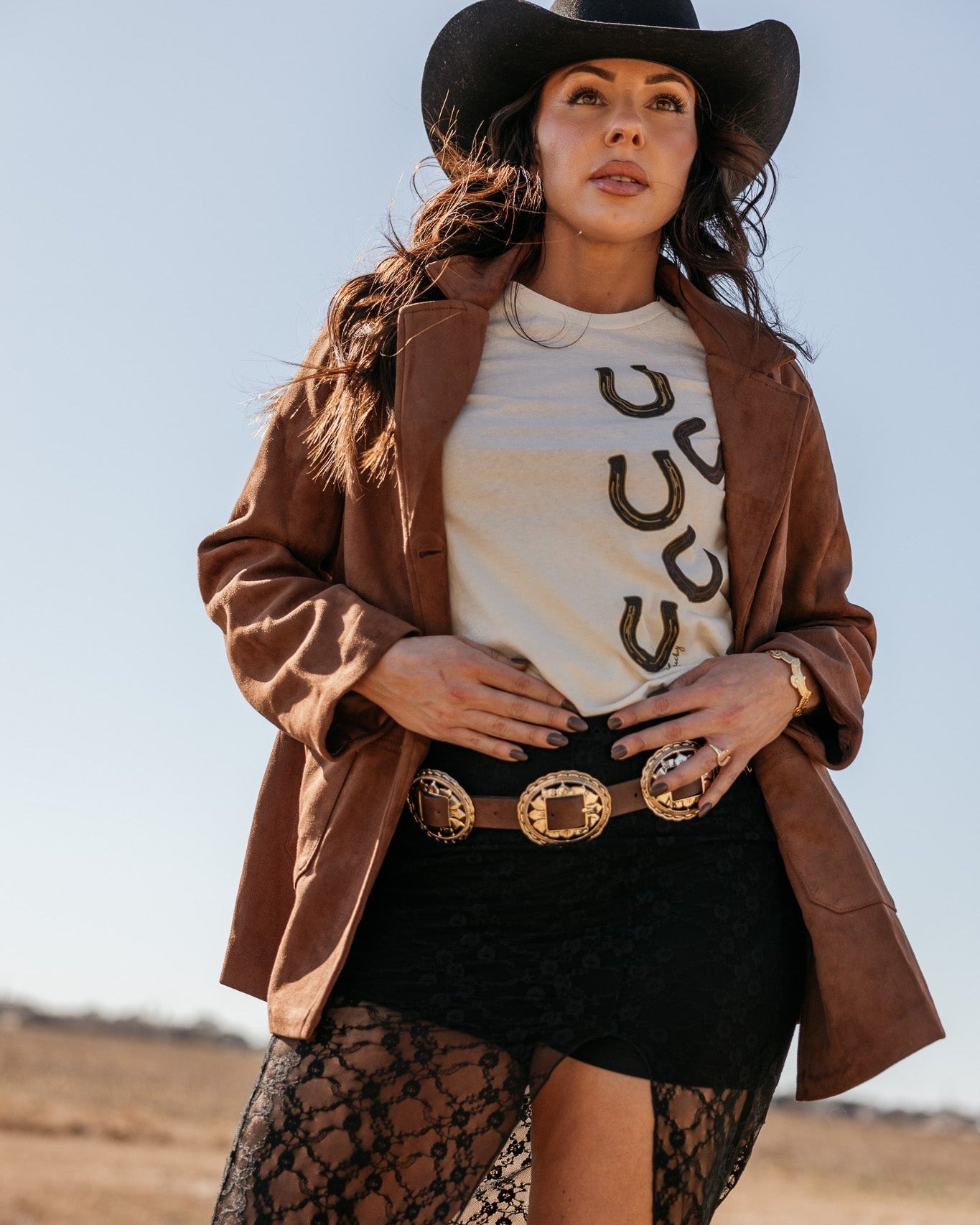Woman in western boho outfit with black cowboy hat, suede jacket, horseshoe tee, and concho belt