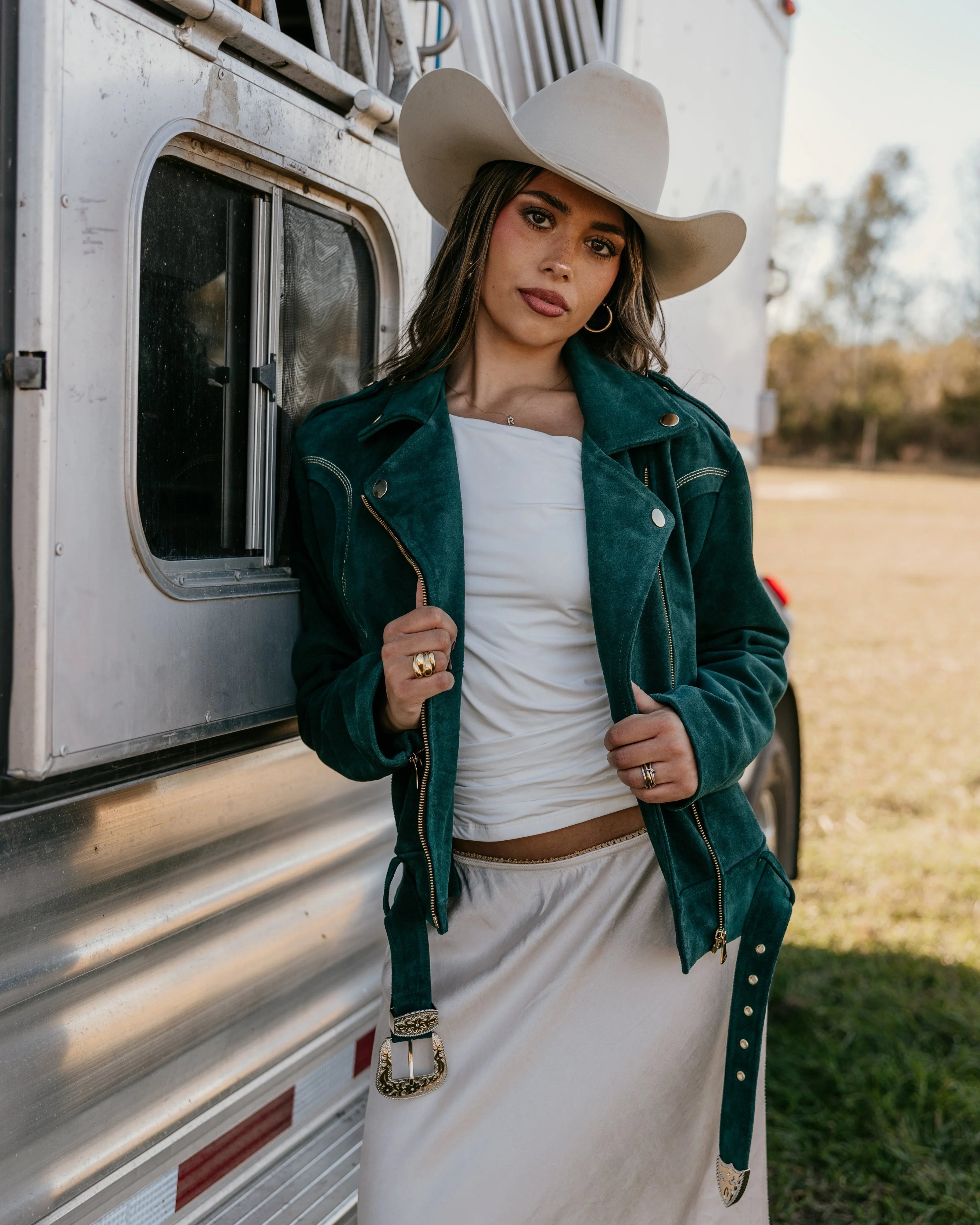 Woman in a suede teal western jacket, white skirt, and cowgirl hat by a horse trailer outdoors