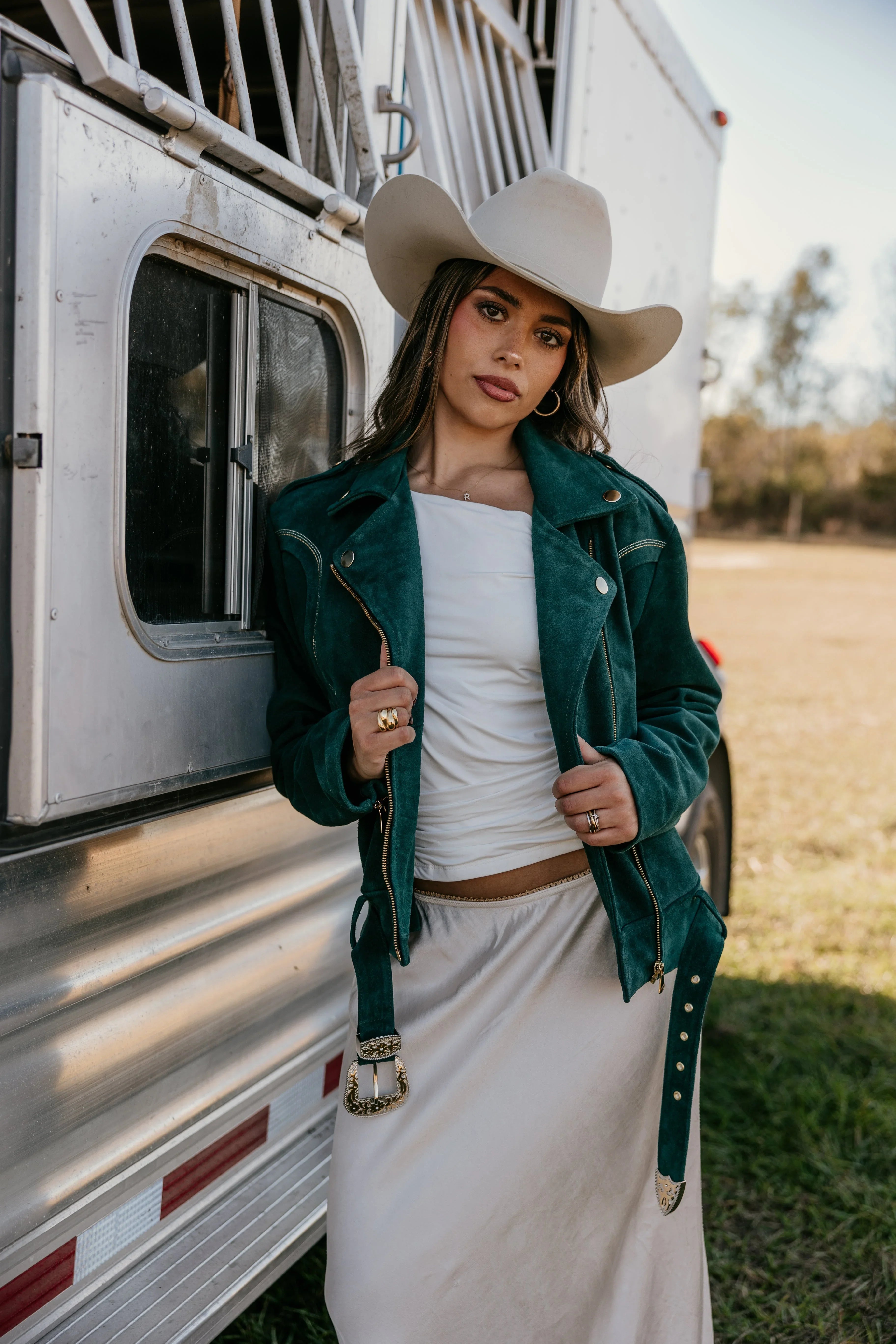 Woman in a suede teal western jacket, white skirt, and cowgirl hat by a horse trailer outdoors