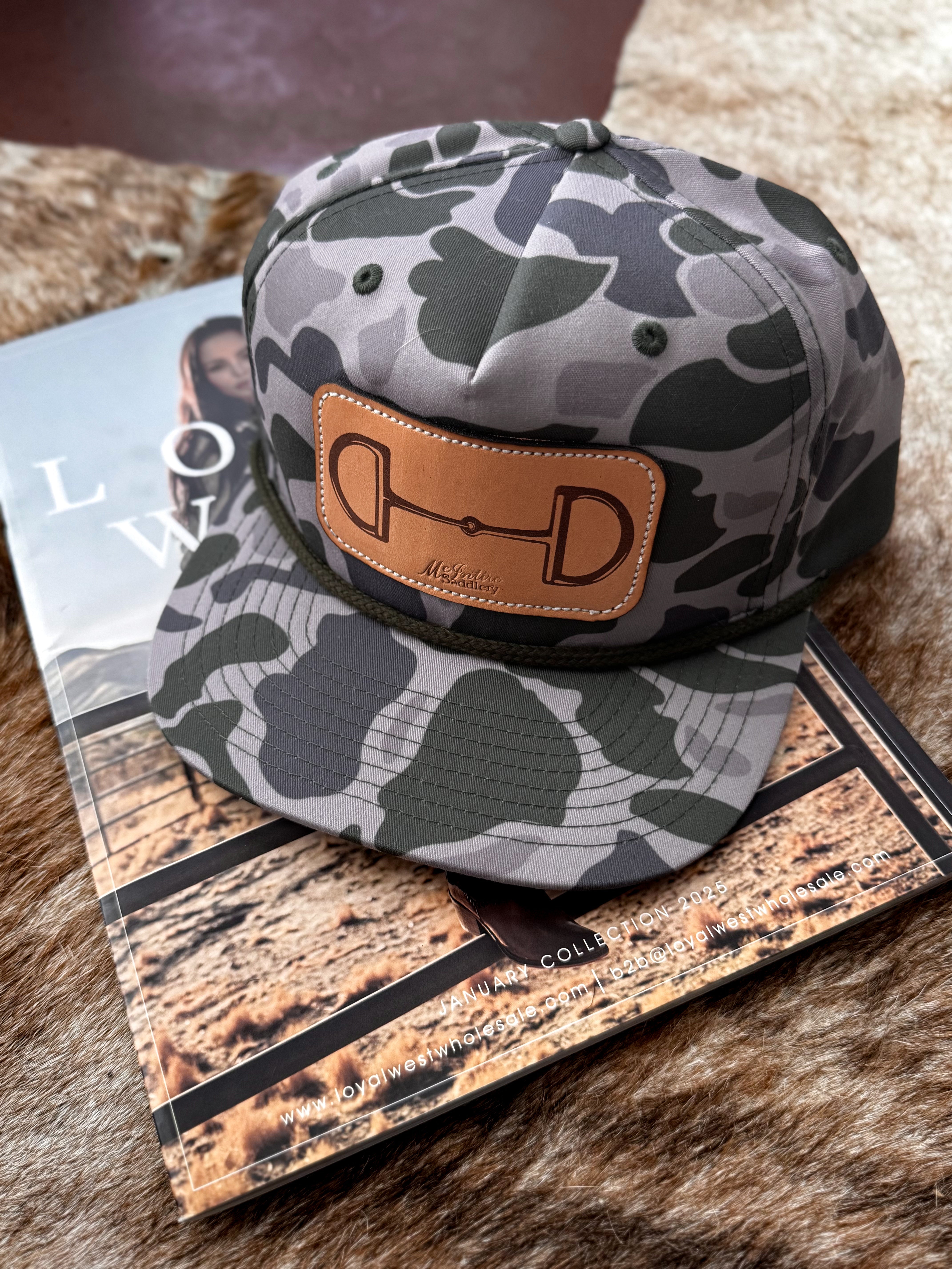 Camo western snapback hat with leather bit patch, styled on a fur surface with a magazine.