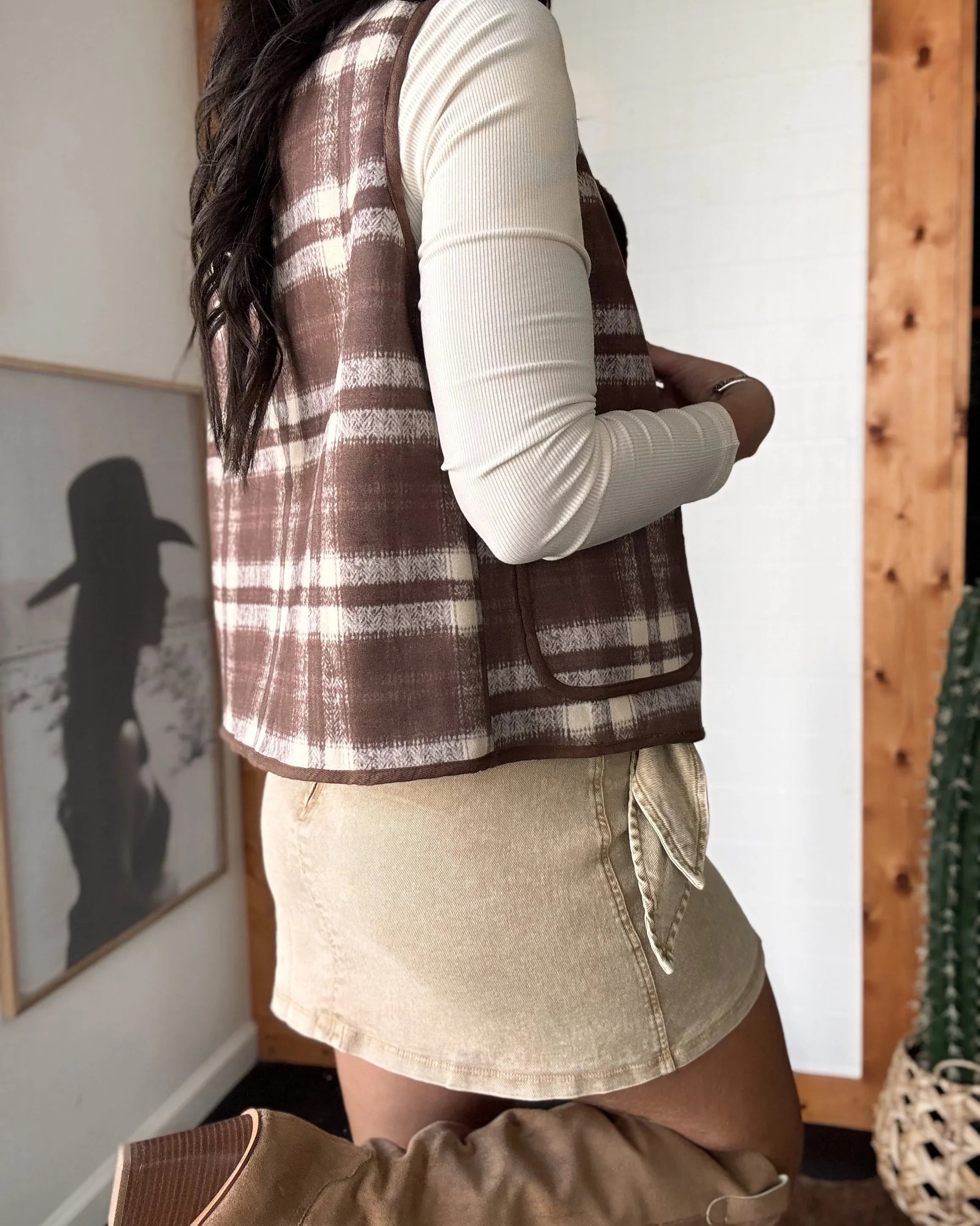 Person wearing a plaid vest over a white long-sleeve shirt and beige skirt in a room with wooden walls.