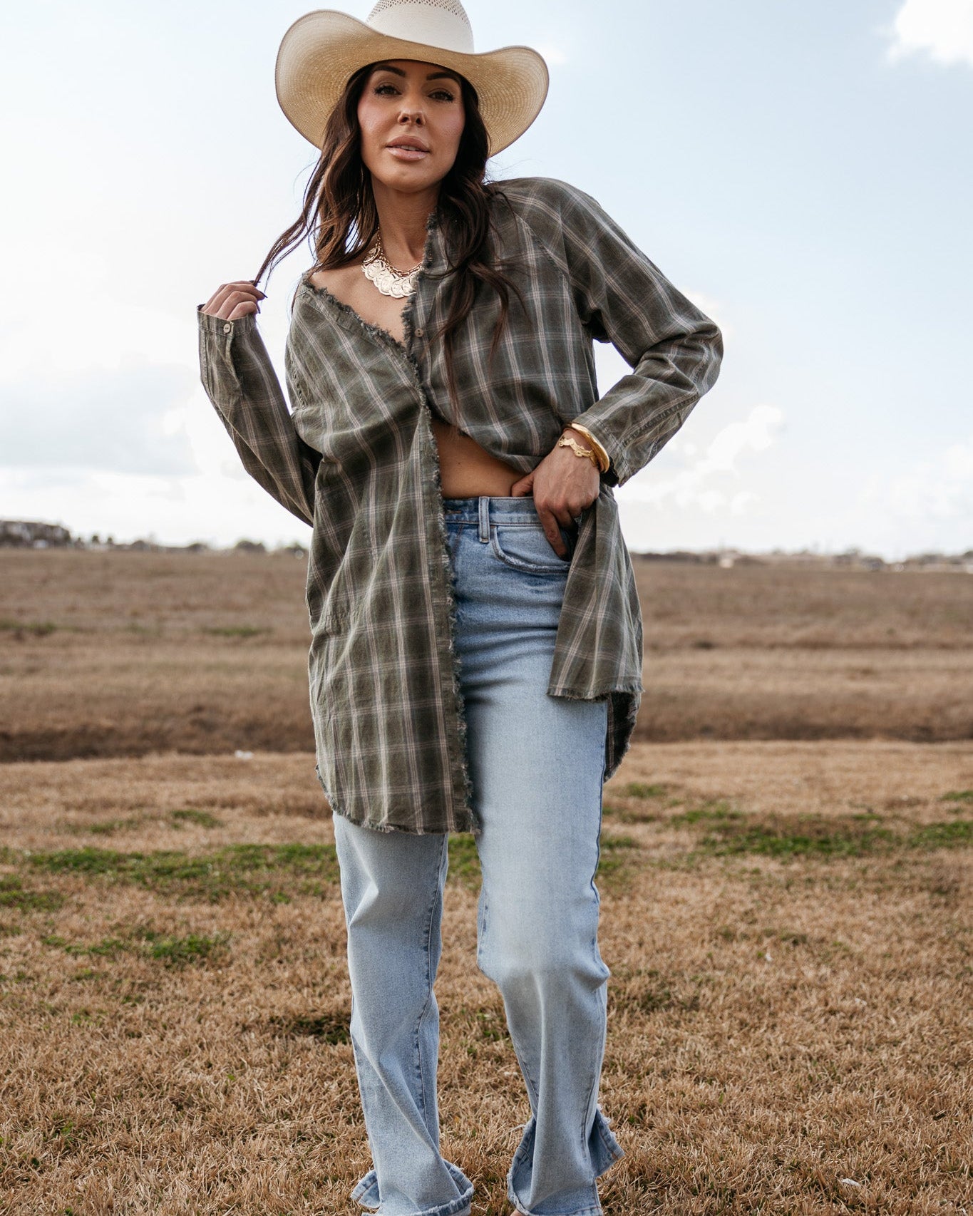Woman in a plaid shirt, cowboy hat, and light wash jeans standing in a western field