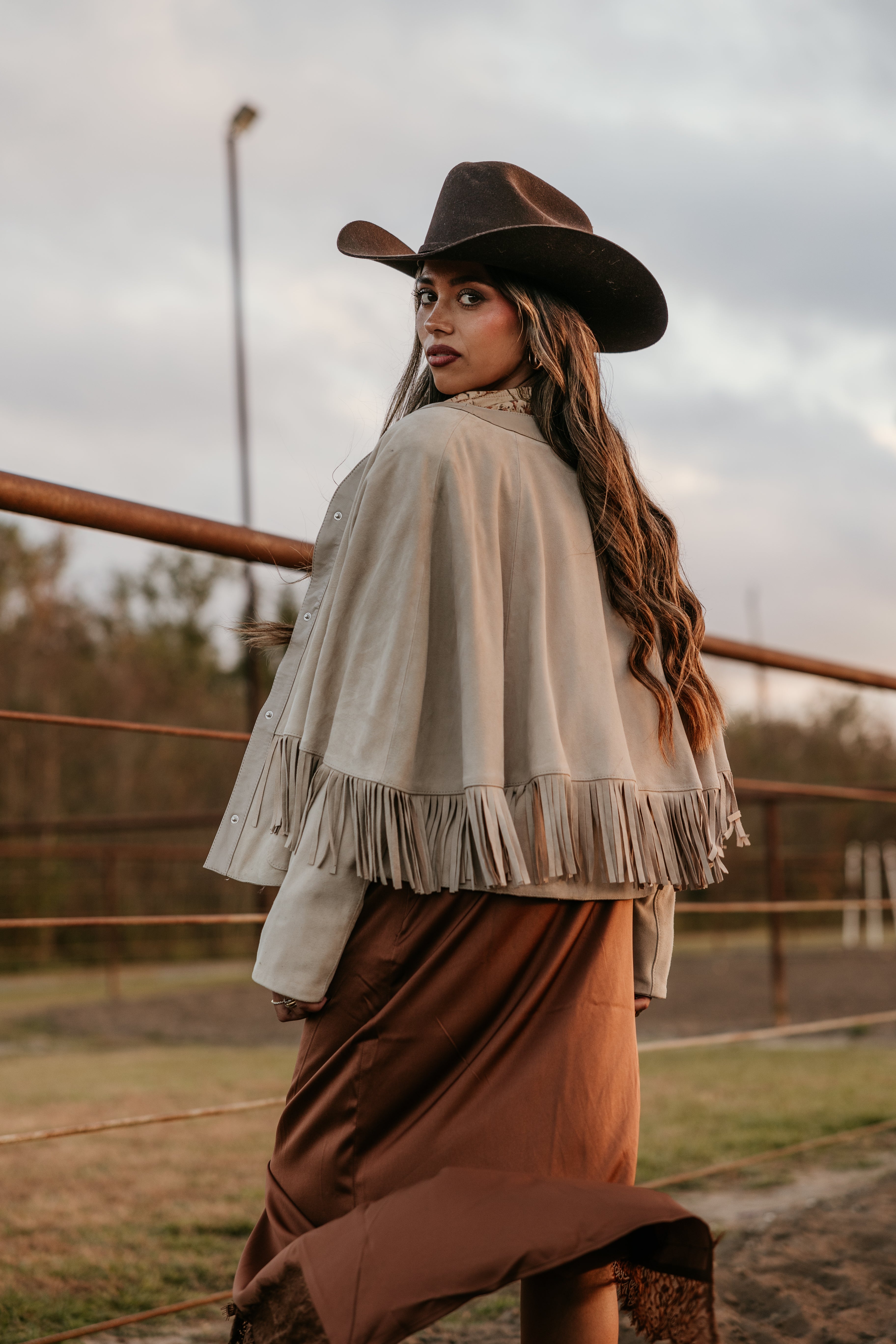 Woman in cowgirl hat and suede fringe jacket at sunset, western boho outfit, outdoor ranch