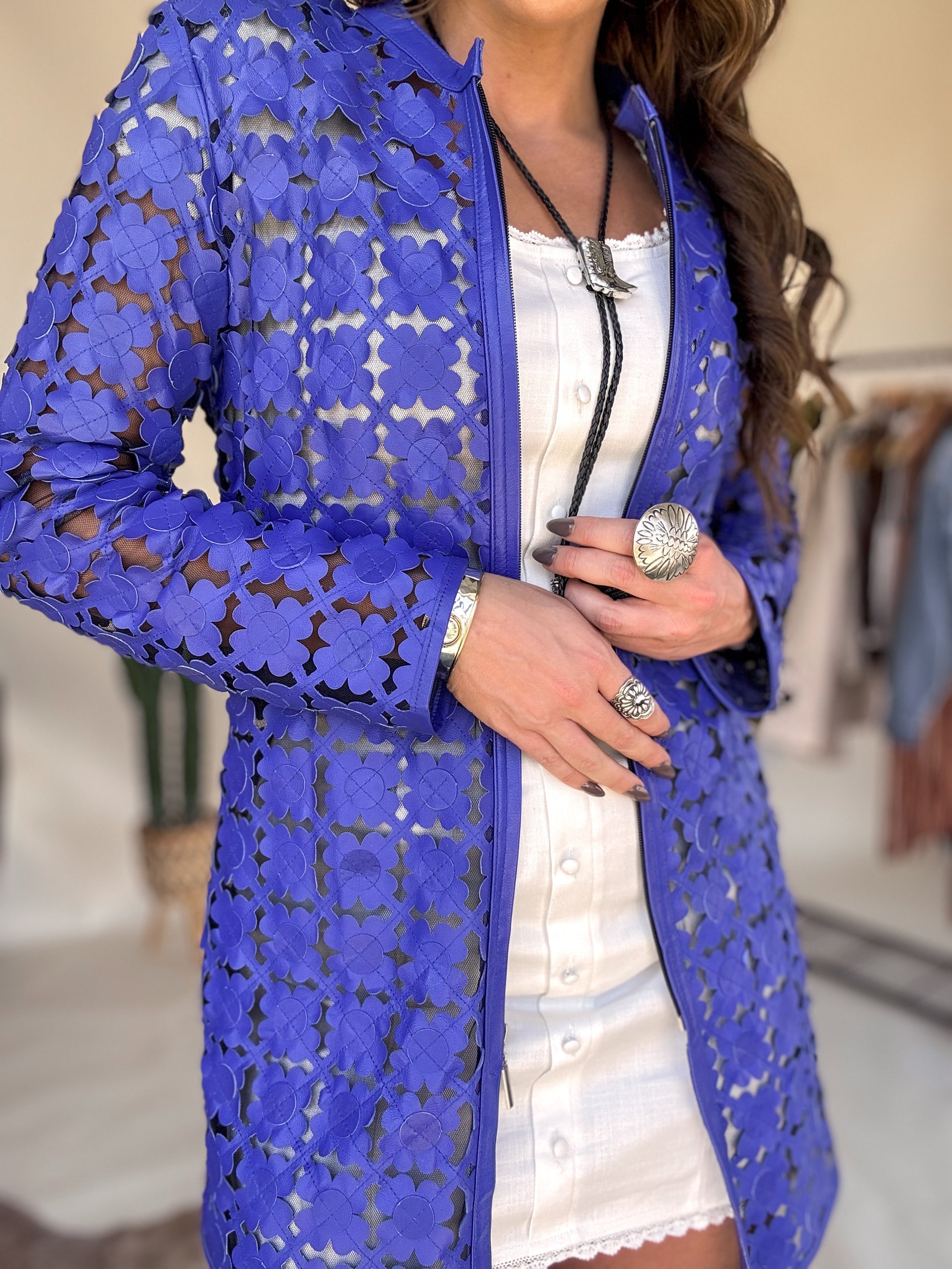 Woman in a white western dress with a bold blue floral mesh jacket, silver jewelry, and bolo tie.