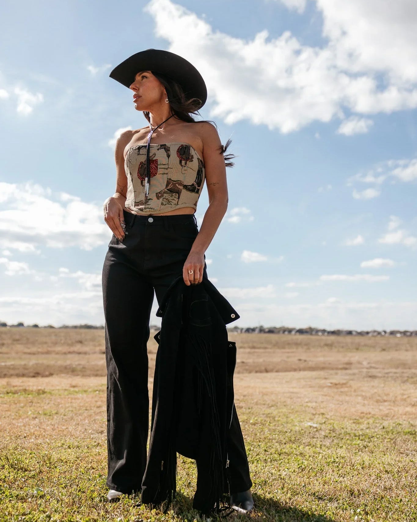 Woman in western boho outfit with black cowboy hat, patterned tube top, and flared pants outdoors.