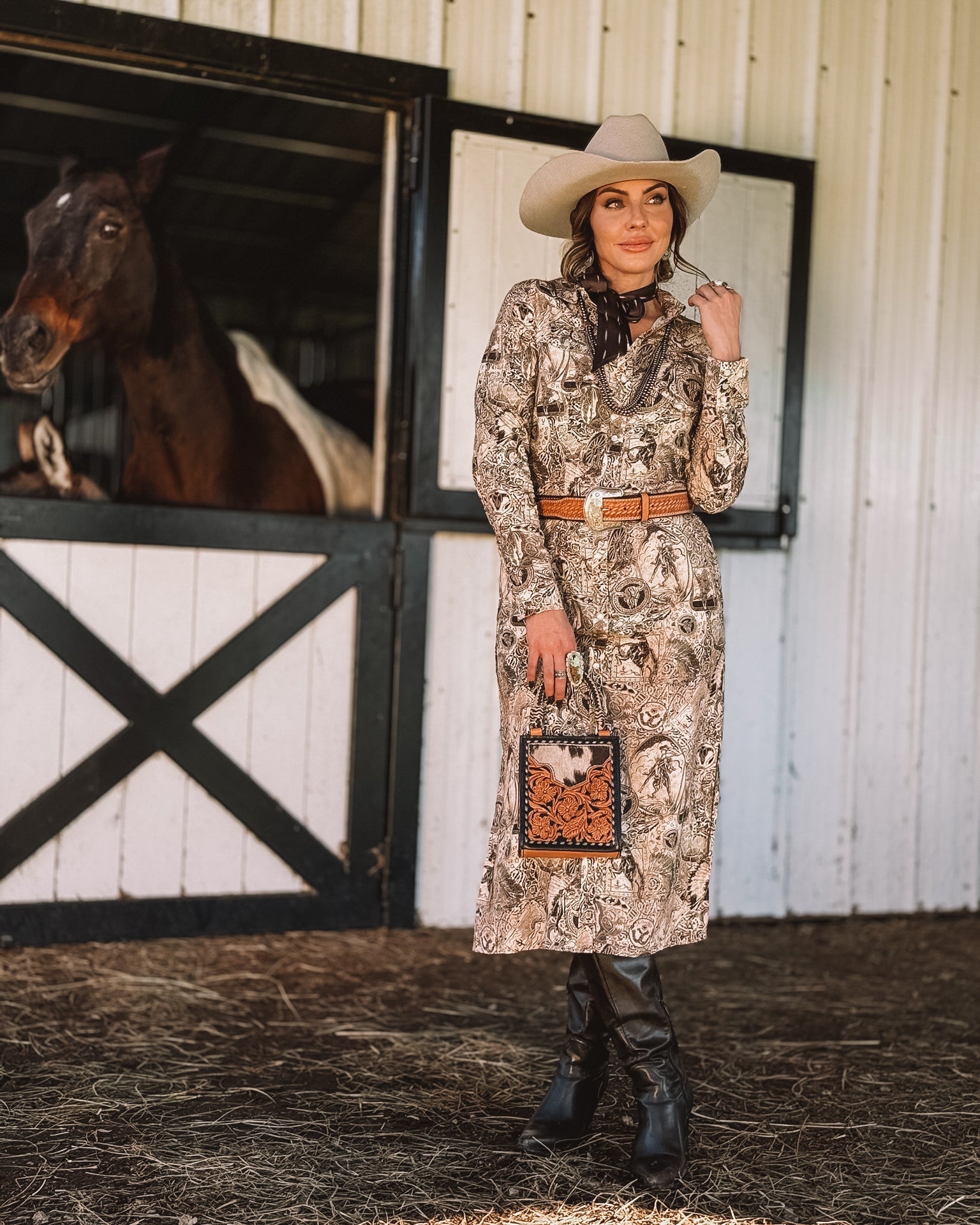 Woman in a western boho dress, cowboy hat, and boots posing by barn with horse for rodeo outfit inspiration