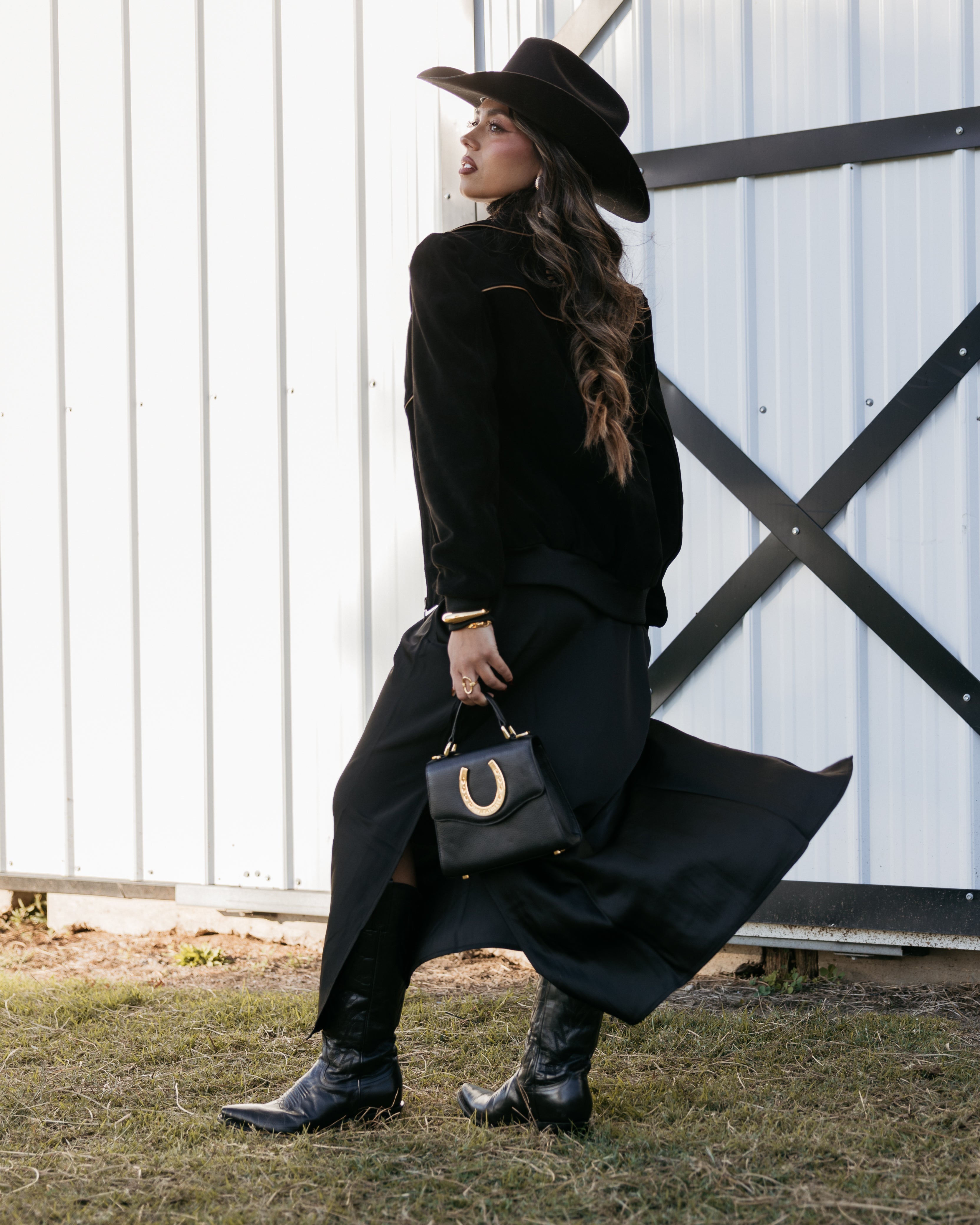 Woman in black cowgirl hat and western outfit with boots and horseshoe handbag outdoors
