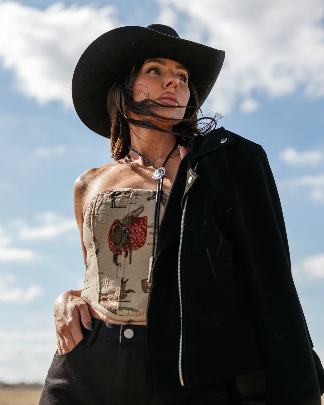 Woman in western boho outfit with black cowboy hat, bolo tie and strapless top against sky