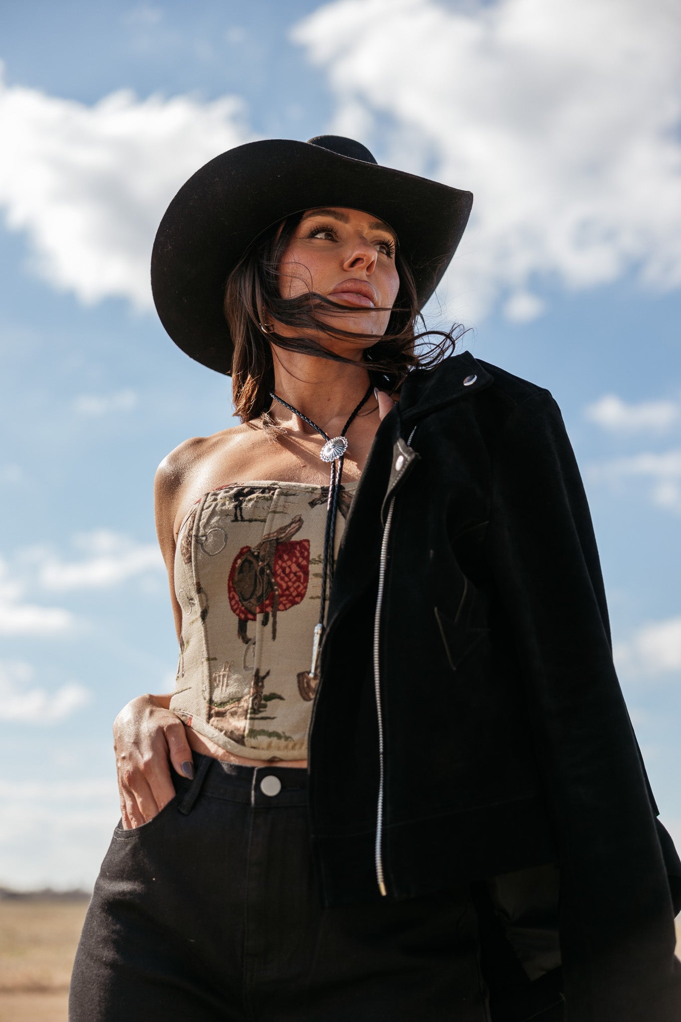 Woman in western boho outfit with black cowboy hat, bolo tie and strapless top against sky