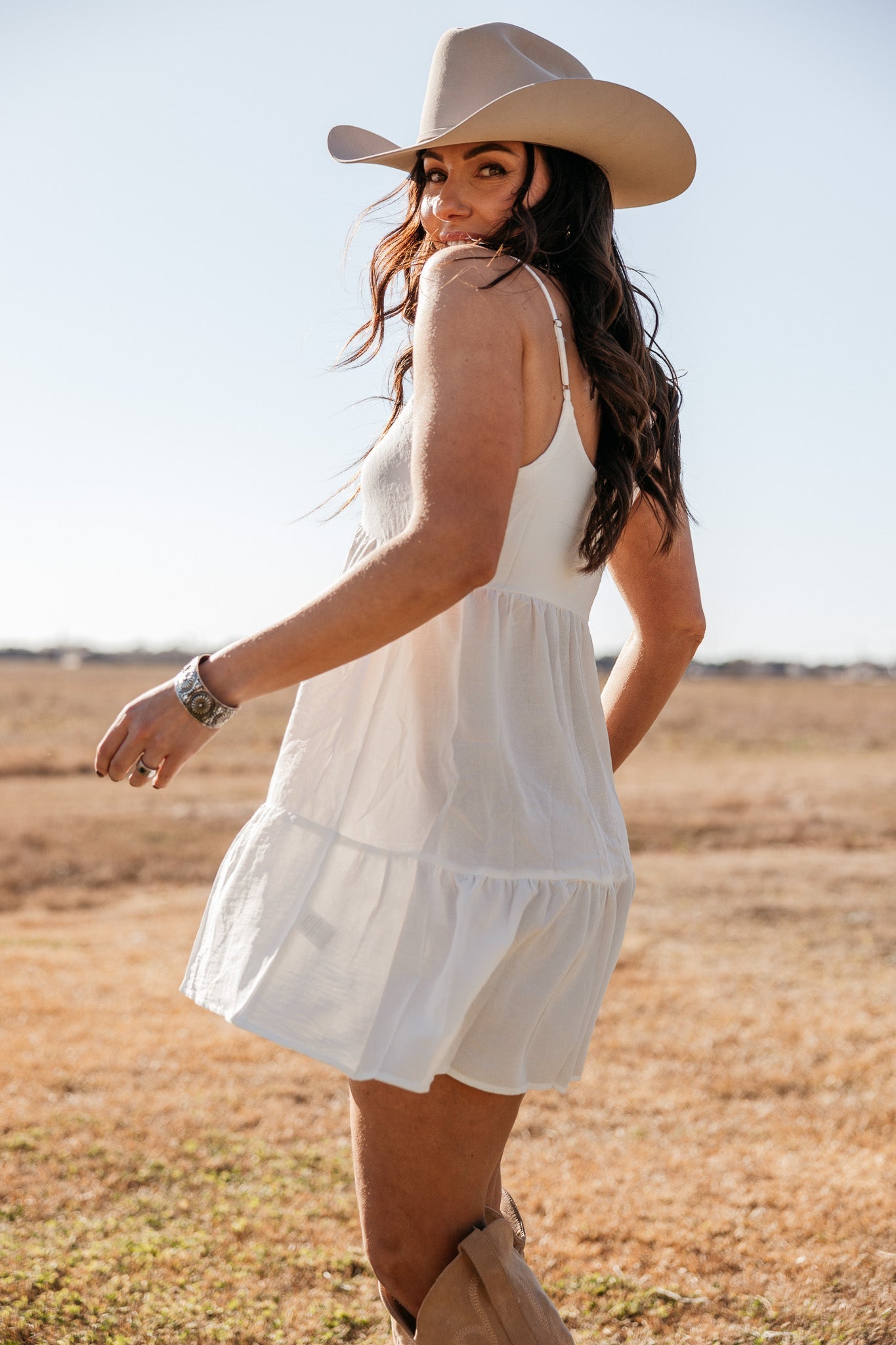 Woman in a white western dress, cowboy hat, and boots outdoors in a boho cowgirl style.