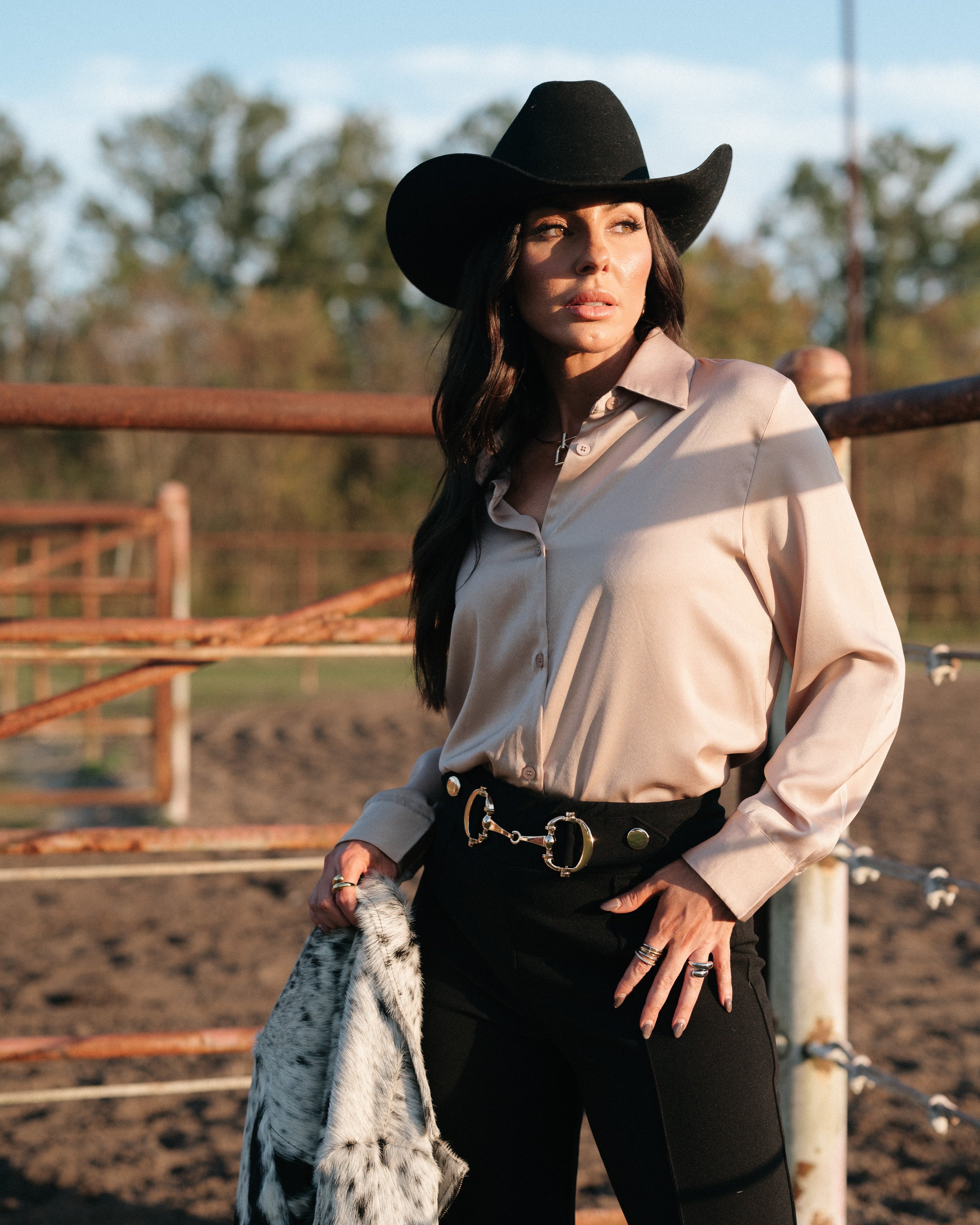 Woman in western boho outfit with black hat, cream blouse, and statement belt by corral fence