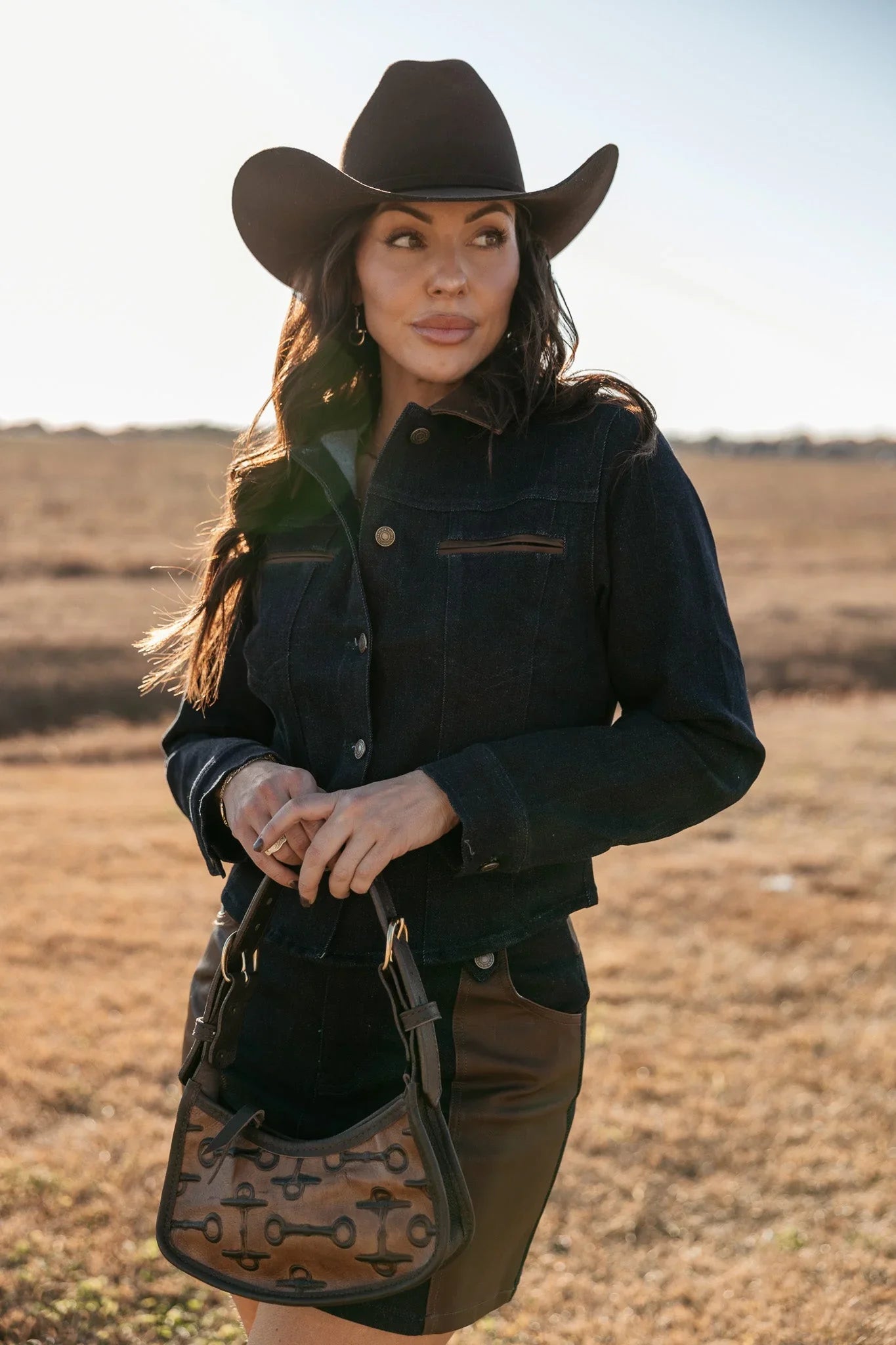 Woman in black cowboy hat and denim jacket with brown purse, western boho style outdoors