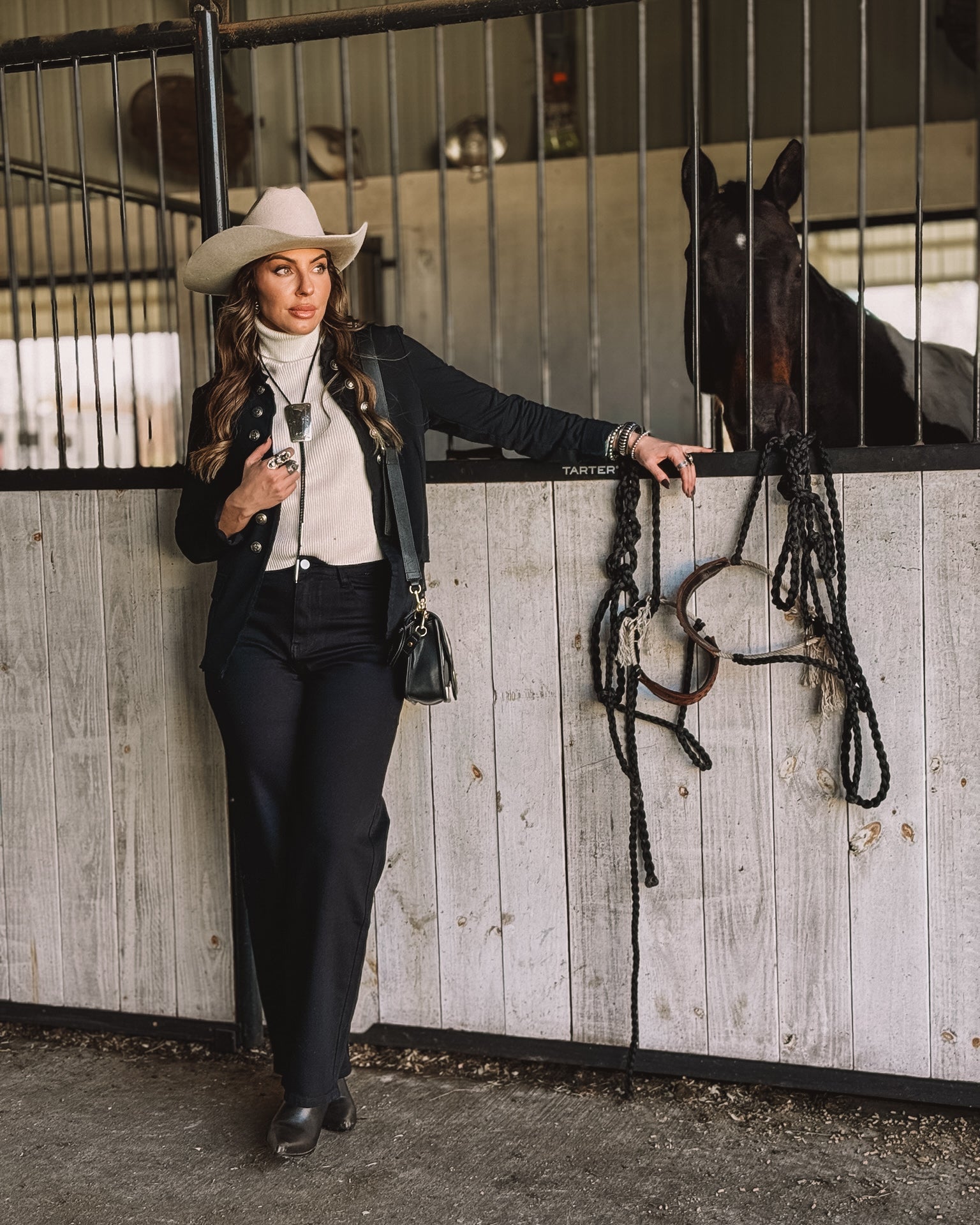 Woman in western outfit with cowboy hat poses by horse stable, showcasing cowgirl style fashion.