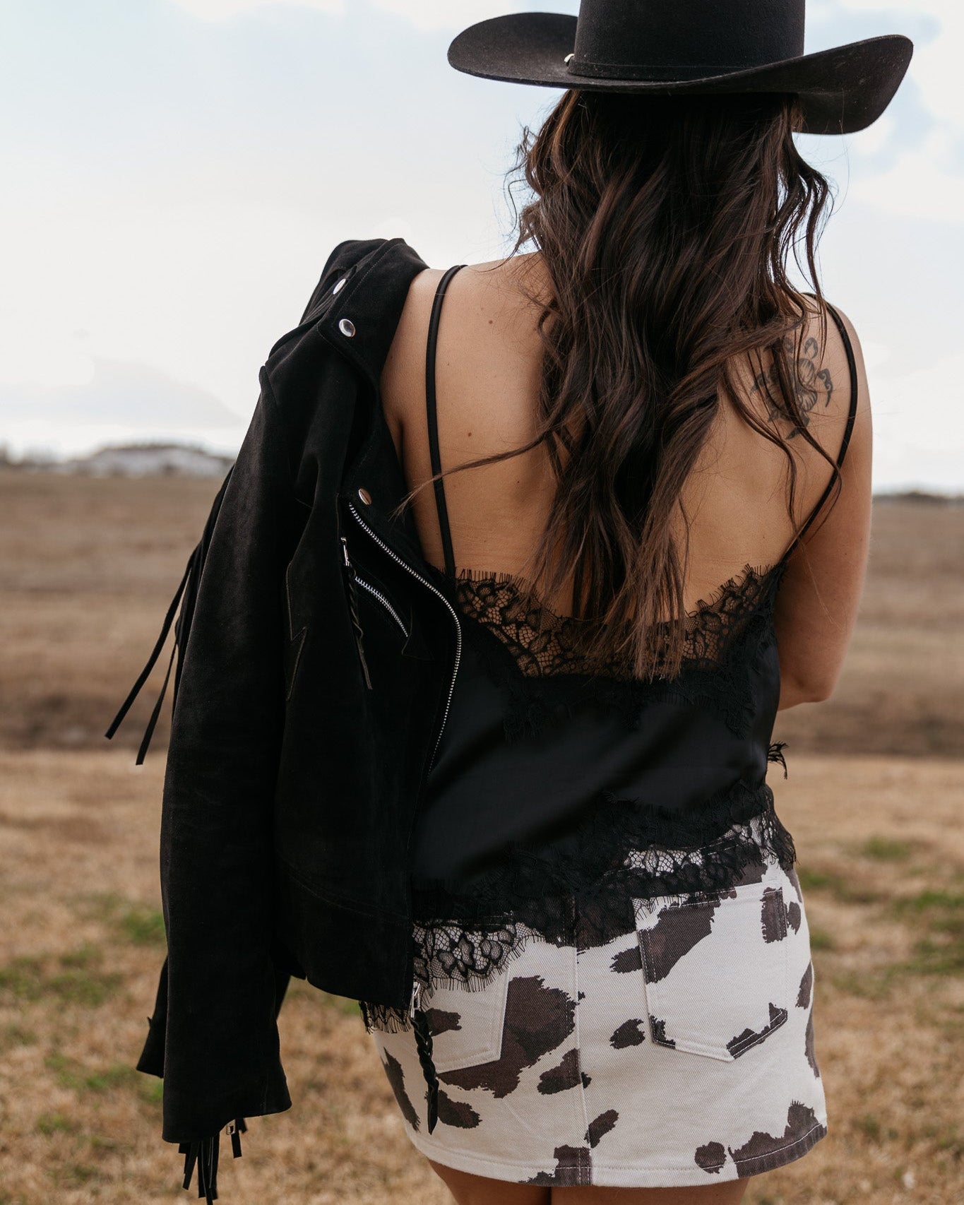 Woman in black cowboy hat, black lace cami, and cow print skirt with suede jacket outdoors