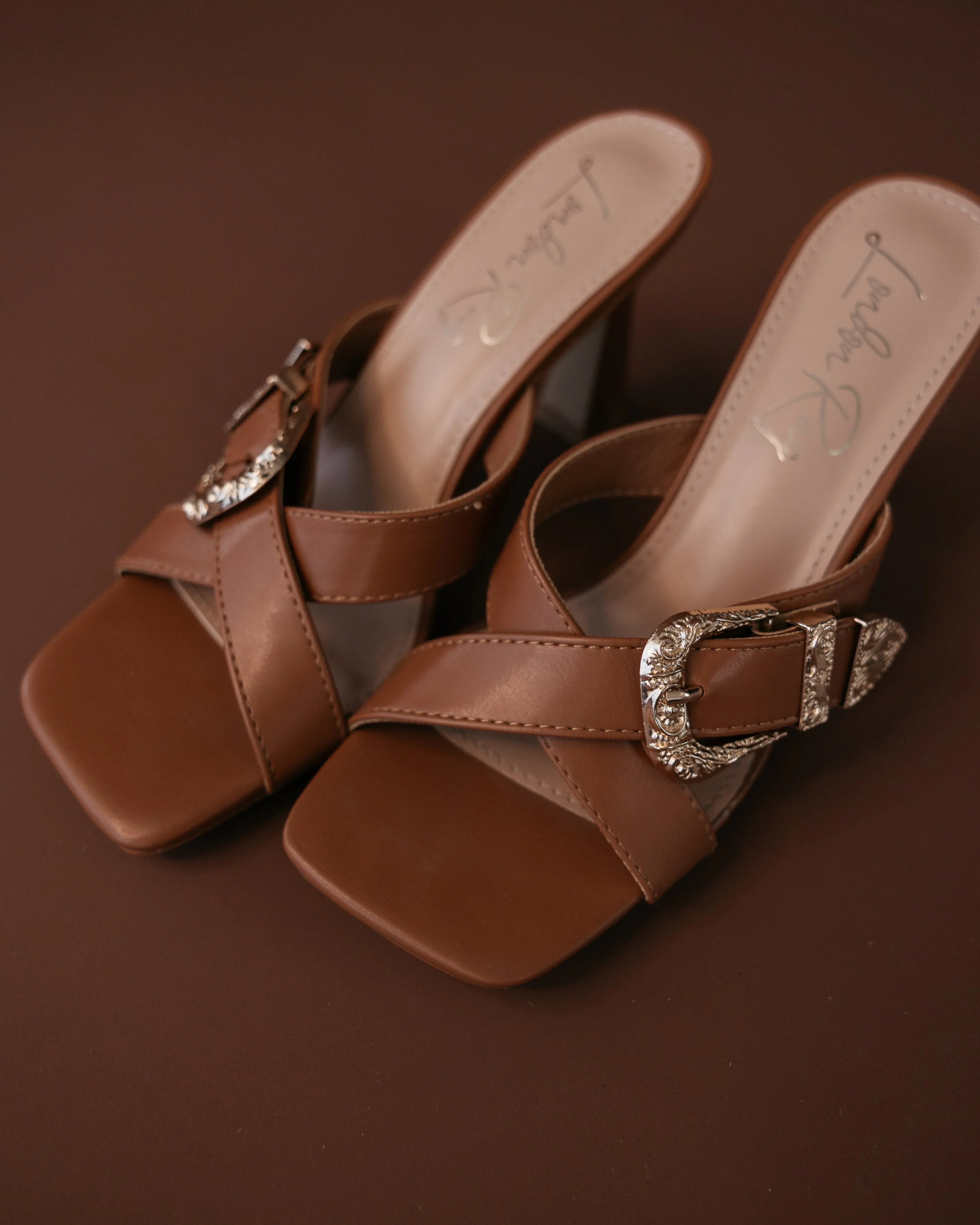 Brown western mule sandals with square toe and silver buckle on brown background