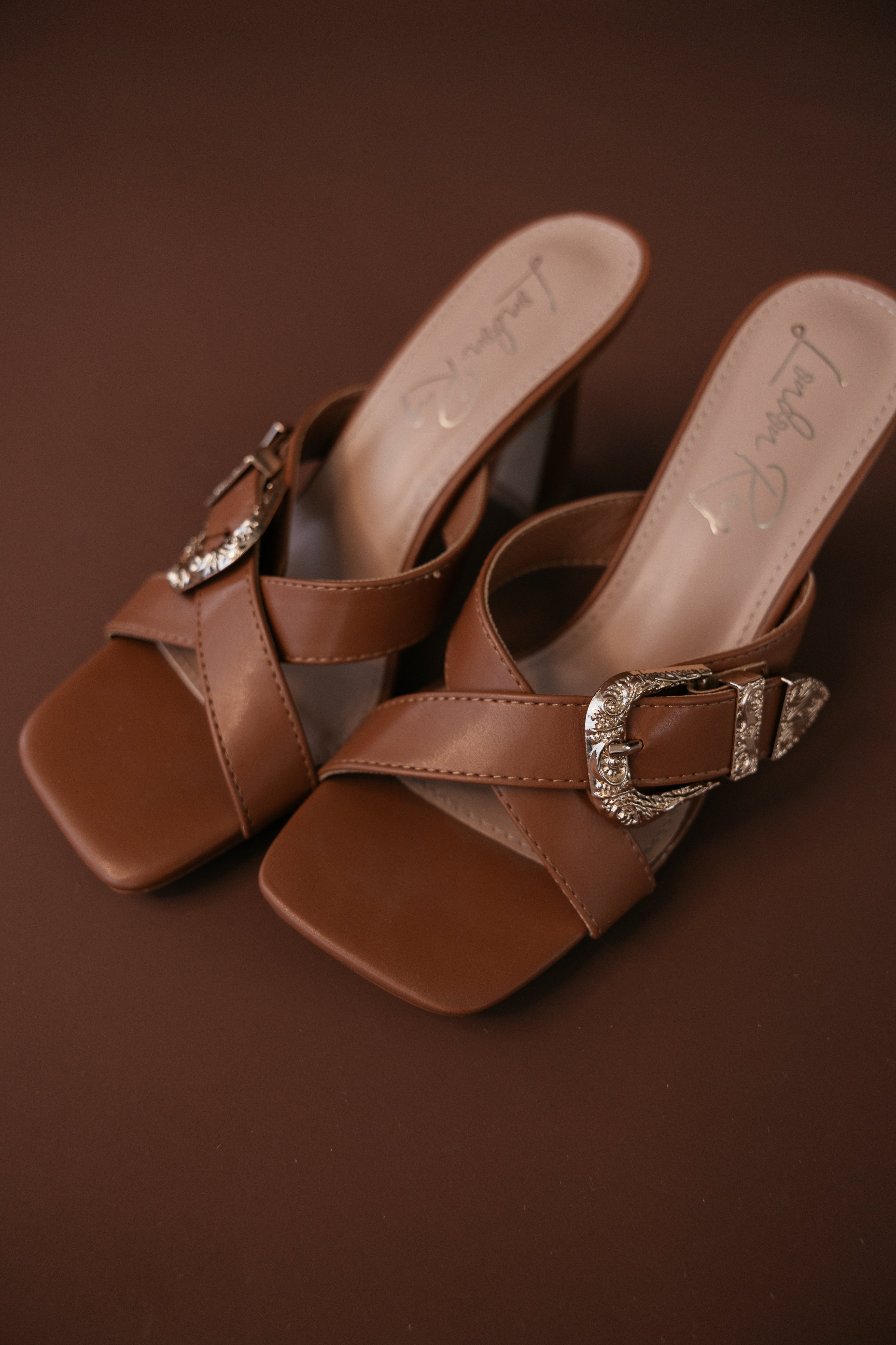 Brown western mule sandals with square toe and silver buckle on brown background
