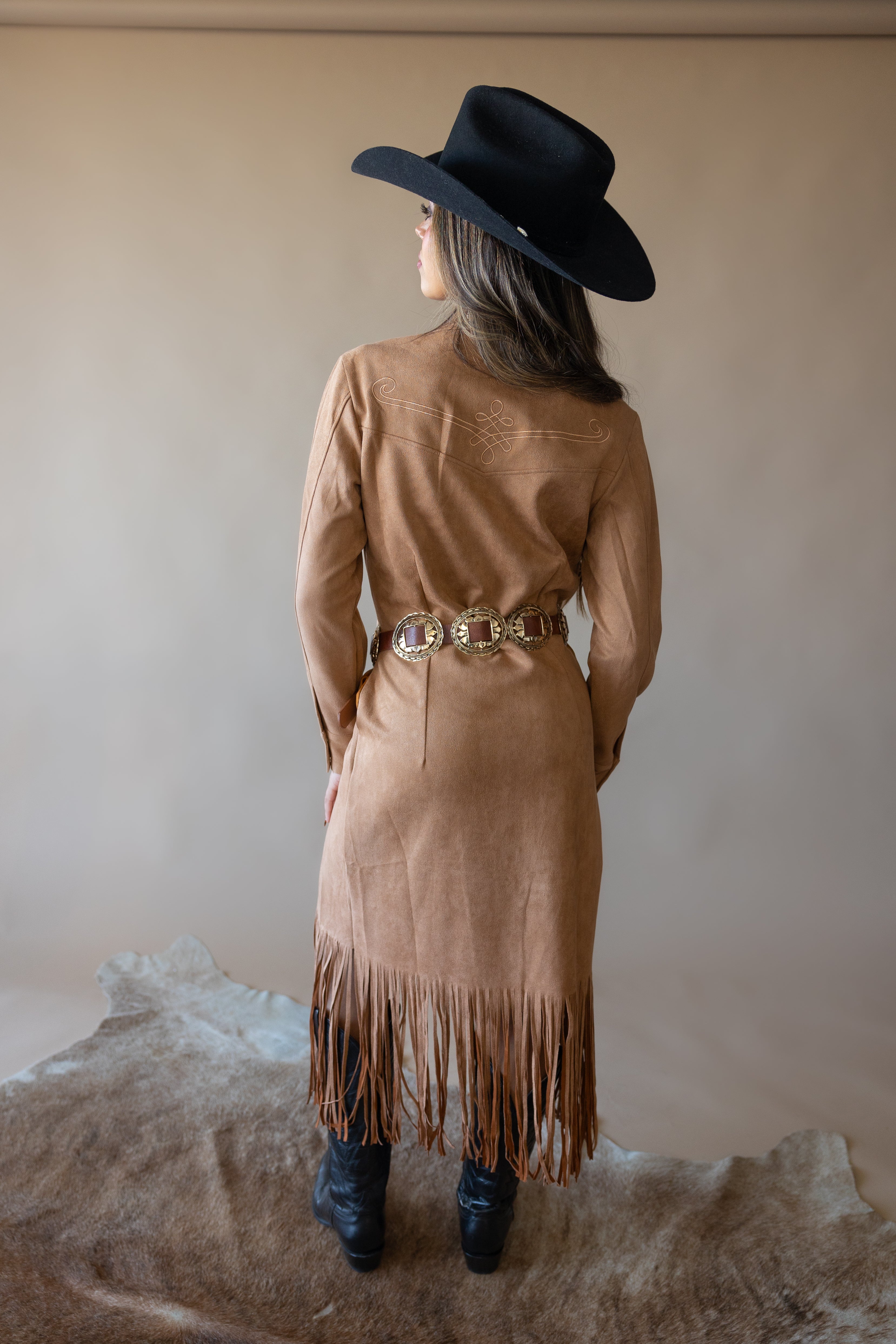 Woman in black cowboy hat, tan fringe suede western dress, and concho belt, back view