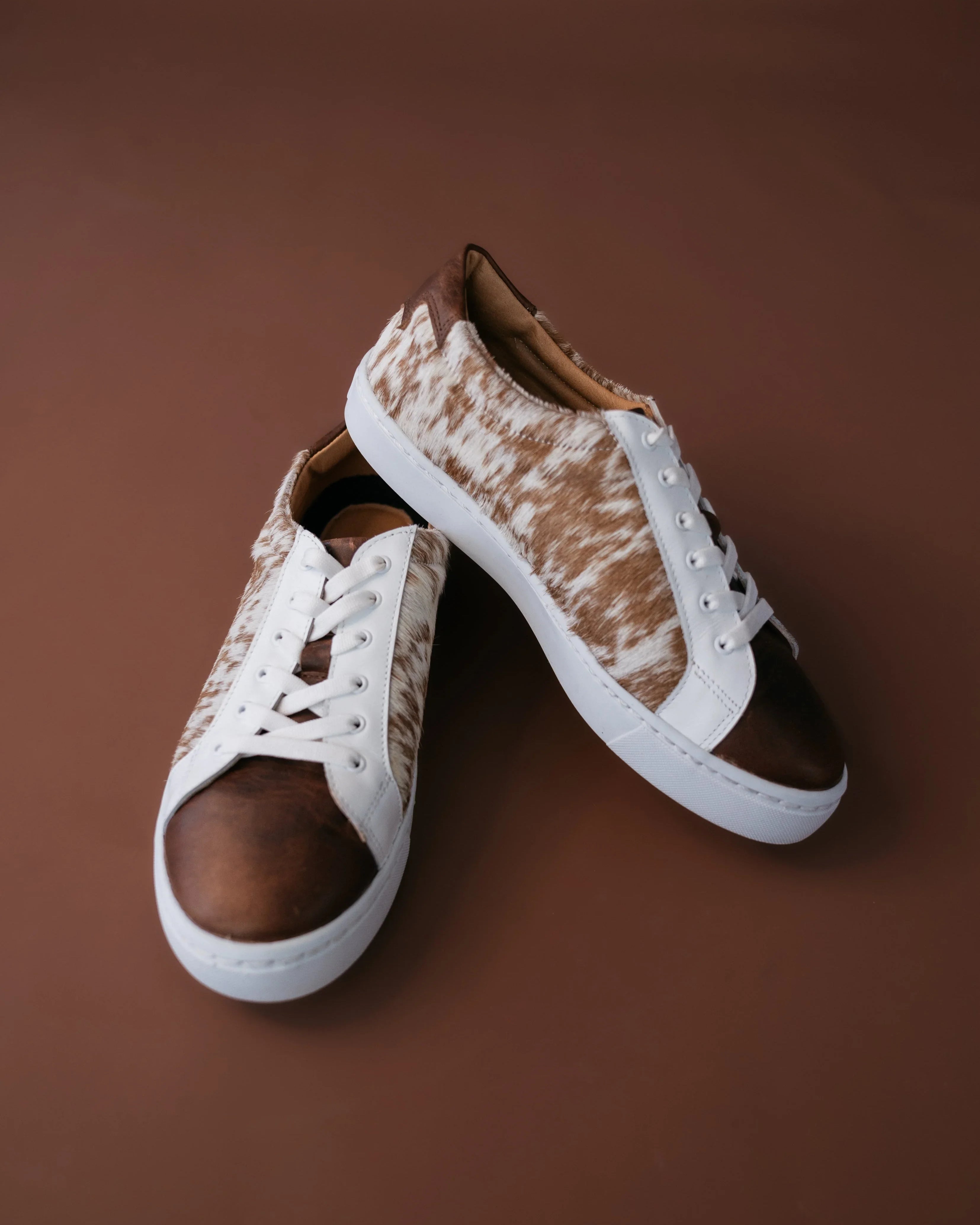 Brown and white cow print western sneakers with white laces on a brown background