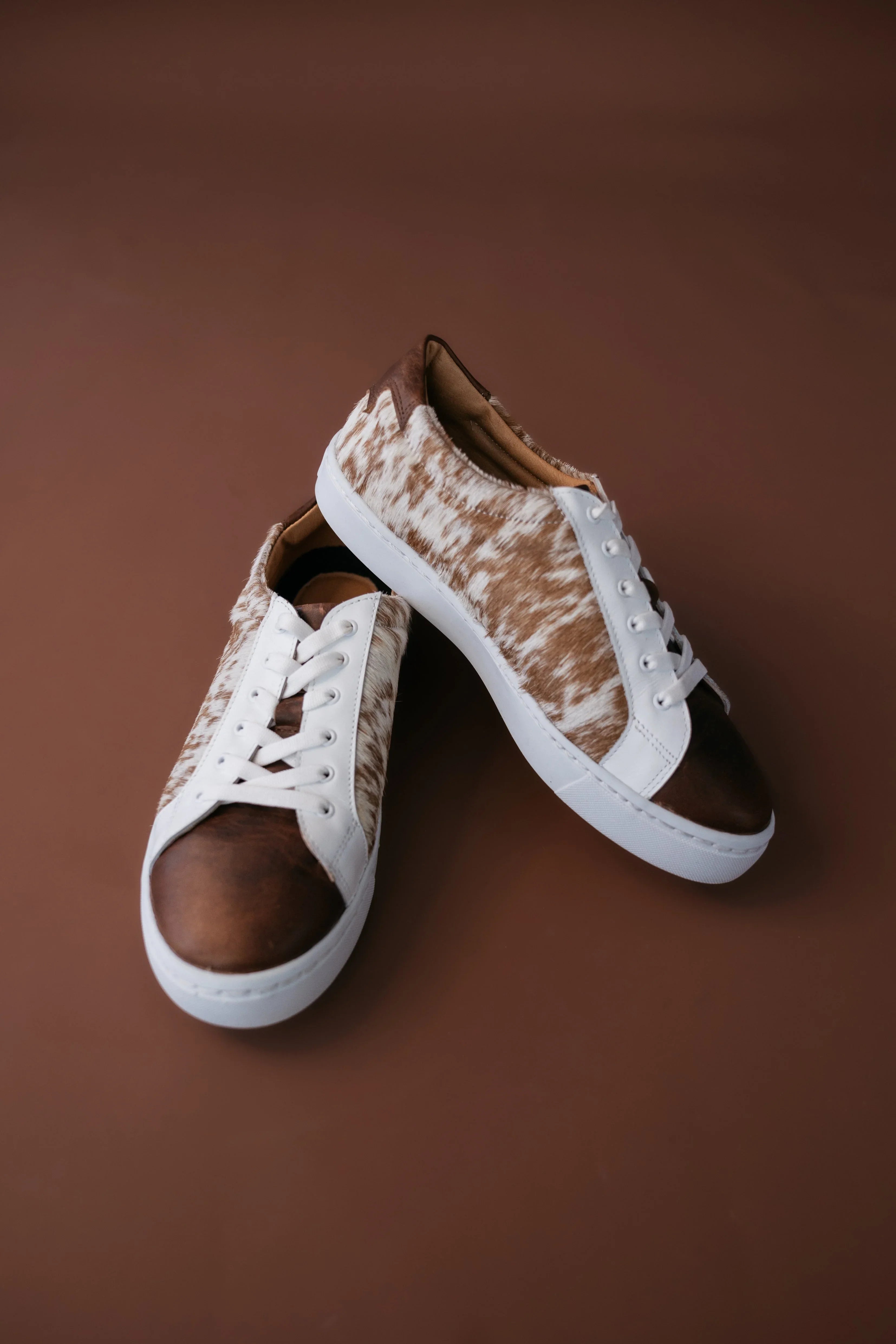 Brown and white cow print western sneakers with white laces on a brown background