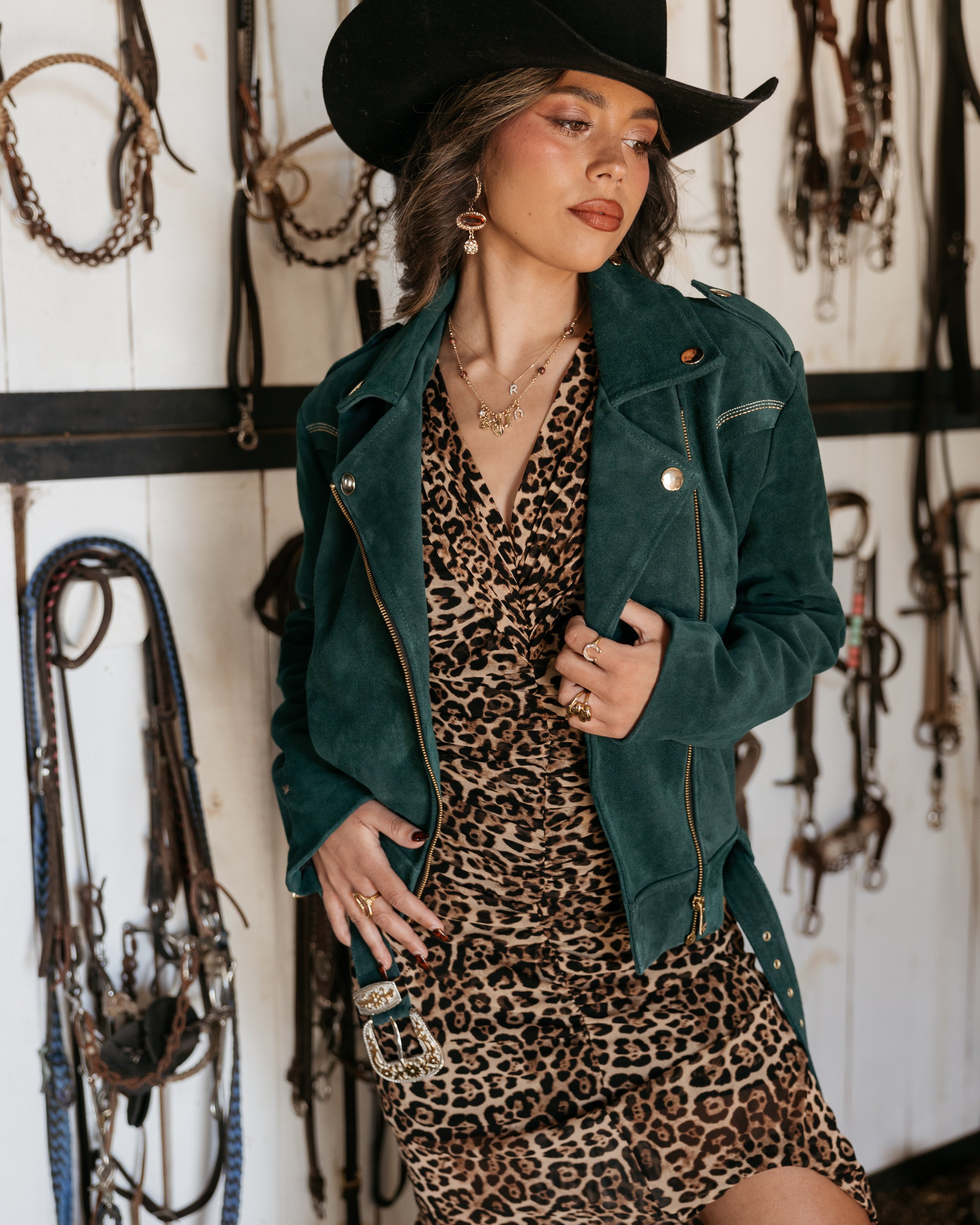 Woman in black cowboy hat, green suede jacket, and leopard print western dress with layered jewelry, standing in a tack room.