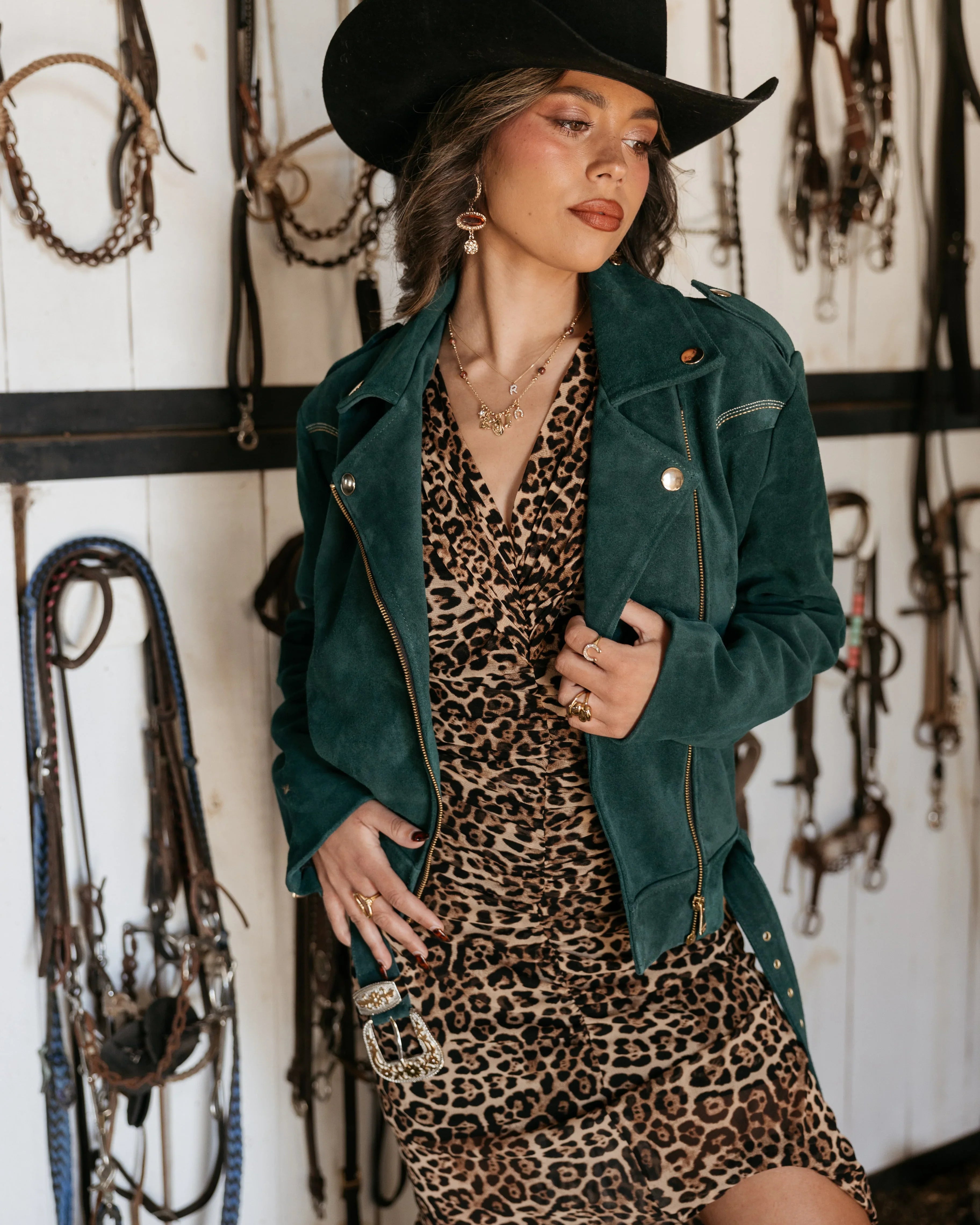 Woman in black cowboy hat, green suede jacket, and leopard print western dress with layered jewelry, standing in a tack room.