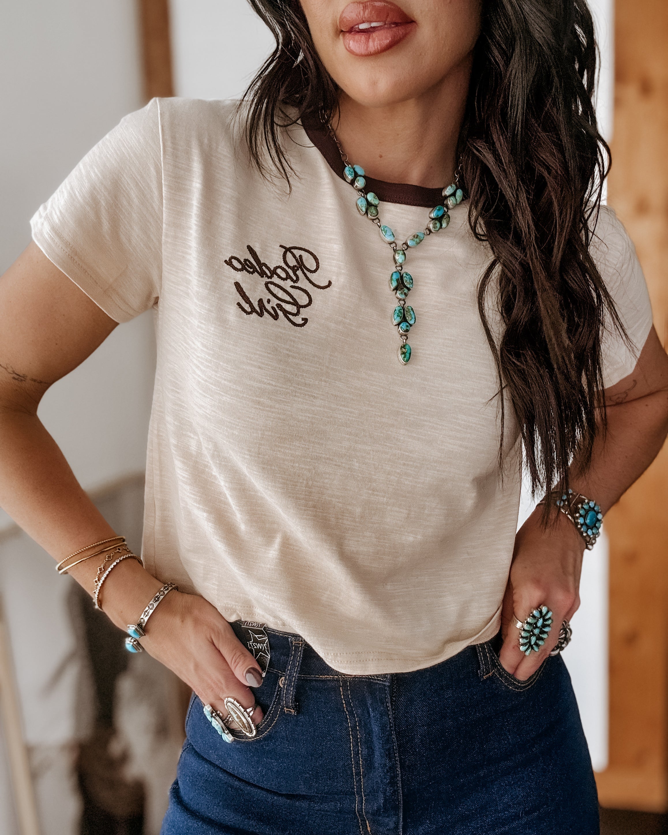 Woman in cream Rodeo Girl t-shirt, turquoise western jewelry, and high-waisted jeans