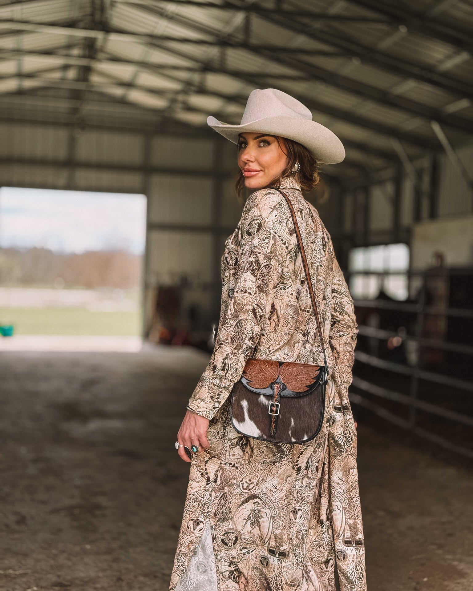 Woman in a western boho dress, cowgirl hat, and cowhide purse in a barn setting.