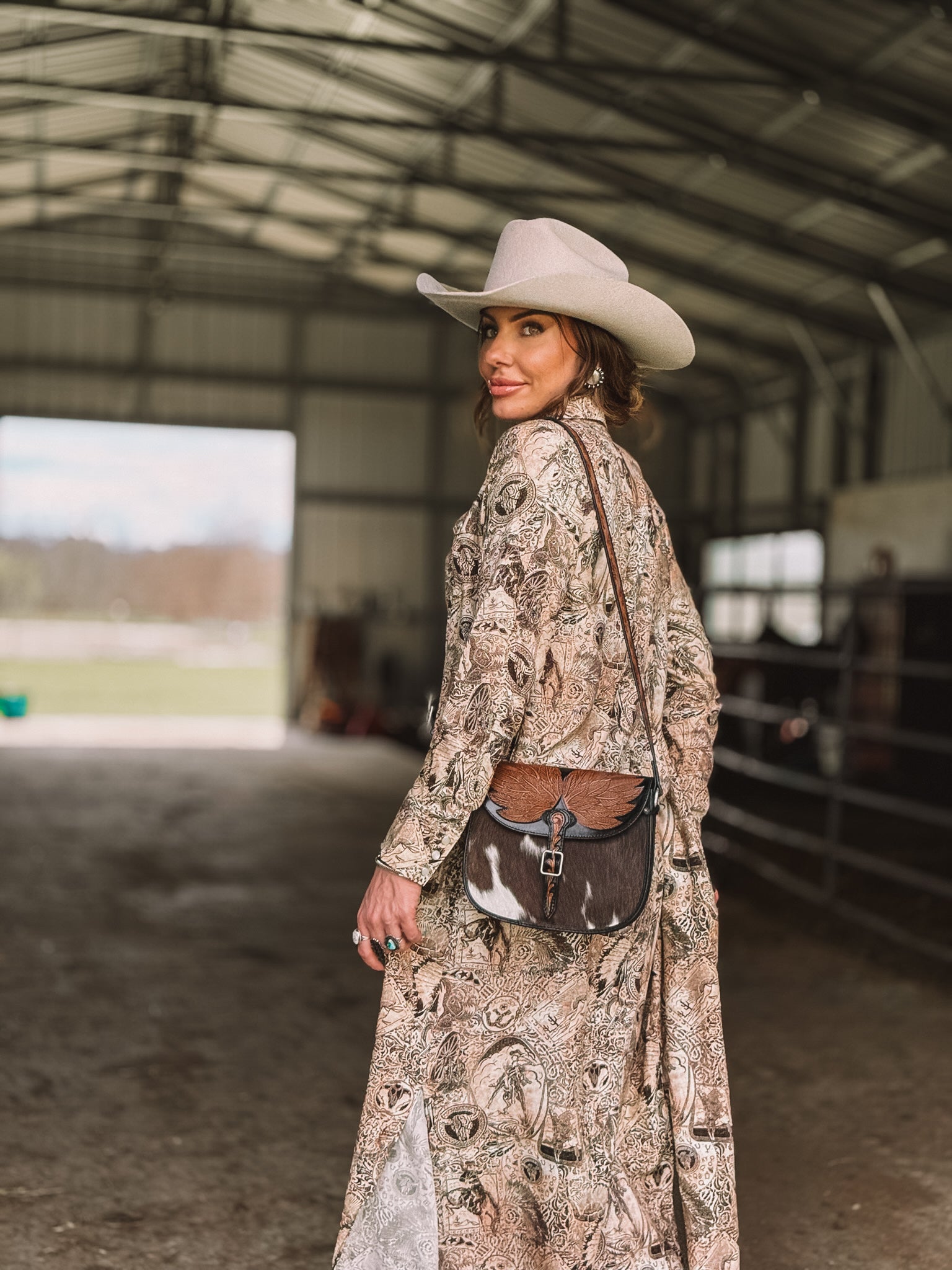 Woman in a western boho dress, cowgirl hat, and cowhide purse in a barn setting.