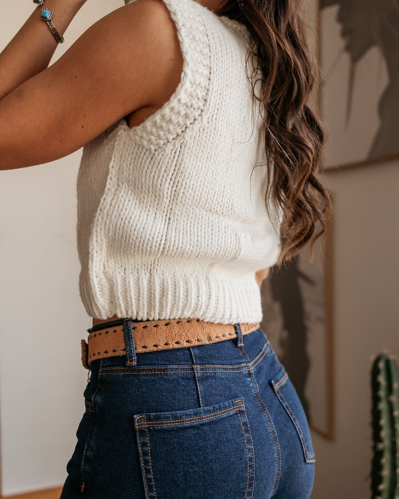 Woman in a white knit sleeveless sweater, blue denim jeans, and tan western belt
