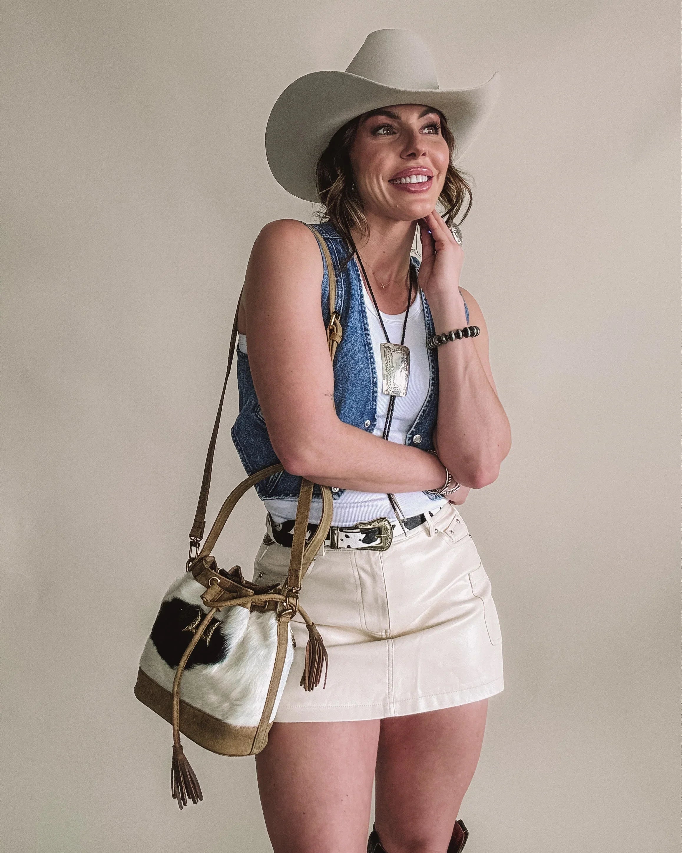 Smiling woman in cowboy hat, denim vest, cream mini skirt, Western bag and jewelry.