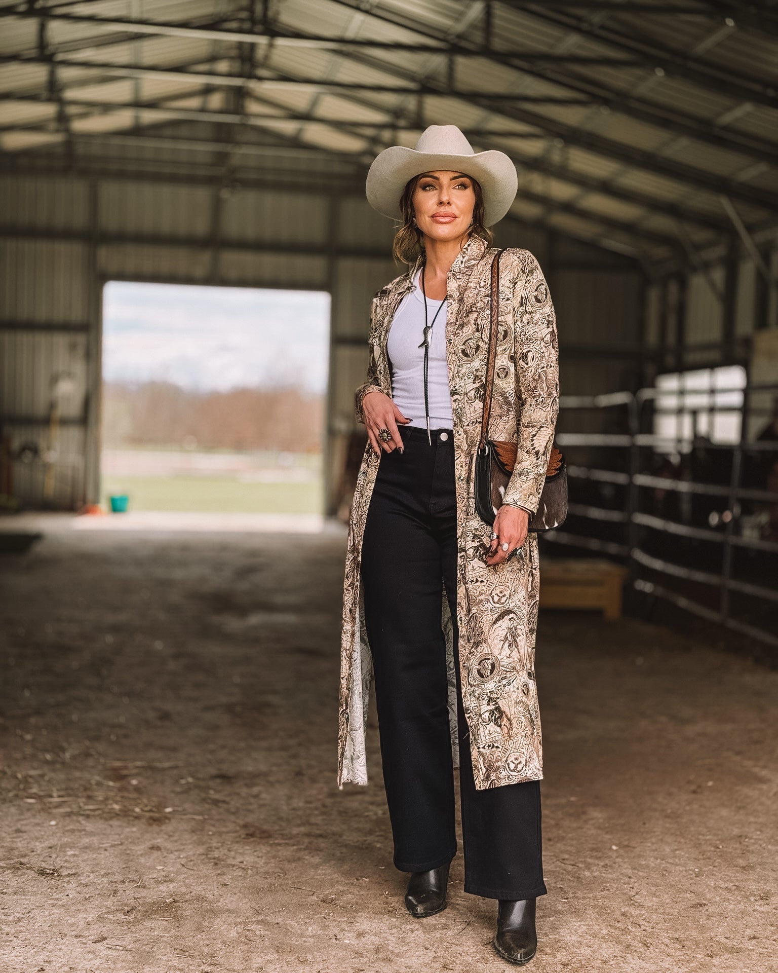 Woman in western boho outfit with paisley duster, black pants, white tank top, and cowboy hat standing in a barn.