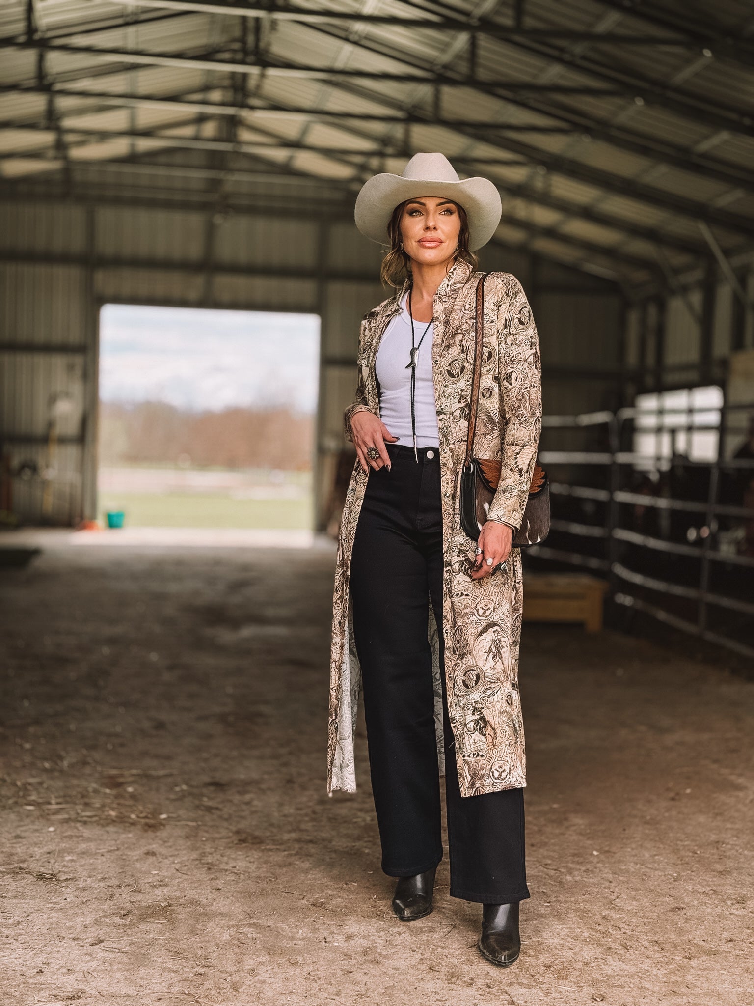 Woman in western boho outfit with paisley duster, black pants, white tank top, and cowboy hat standing in a barn.