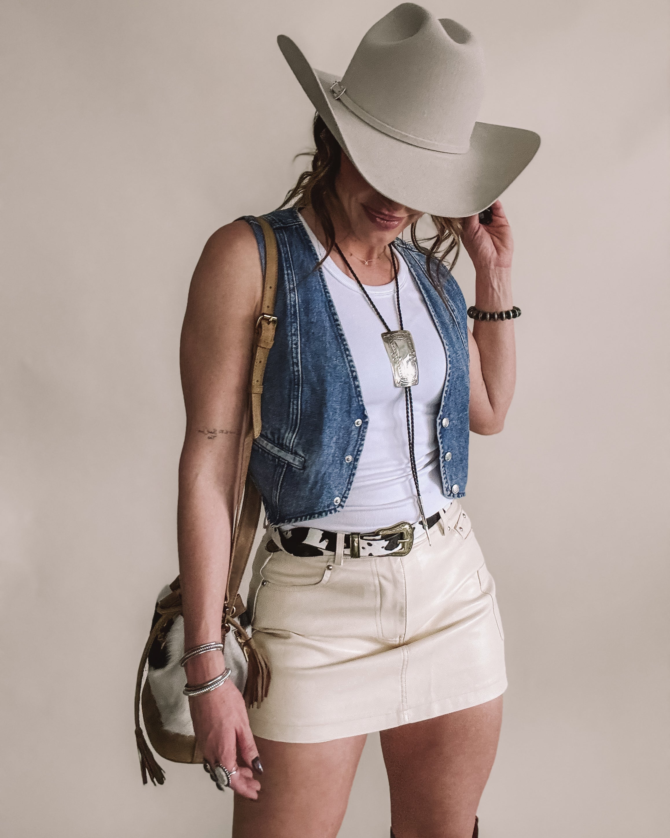 Woman in a western cowgirl outfit with felt hat, denim vest, cream mini skirt, and cow print belt