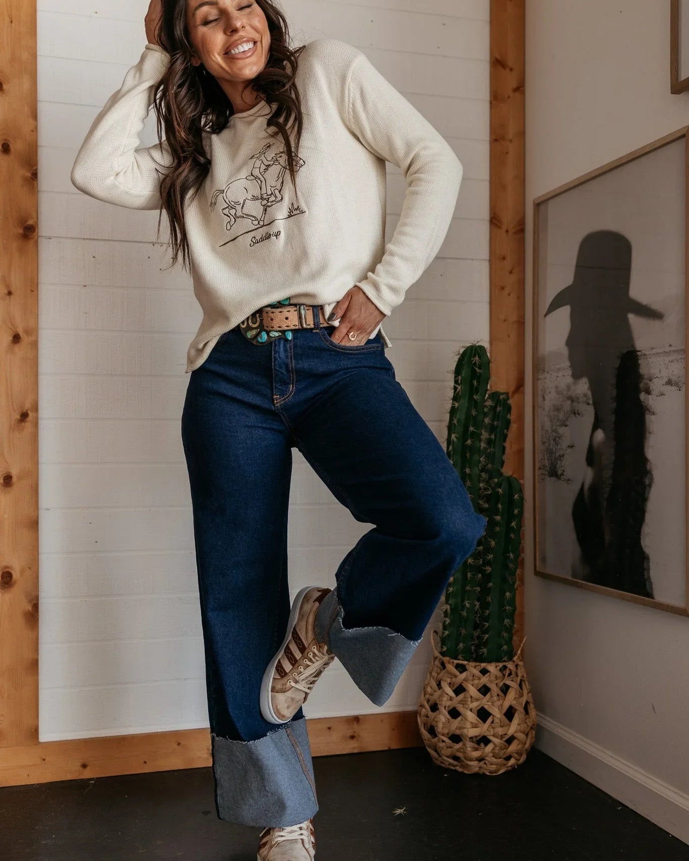 Woman in western boho outfit wearing graphic saddle up sweater and wide-leg jeans indoors
