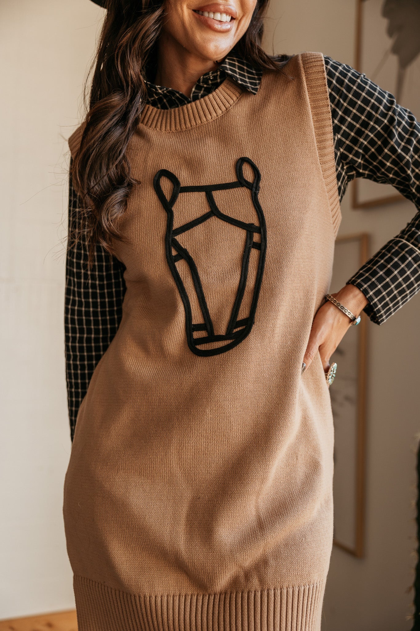 Woman wearing a tan western sweater vest with black horse head outline over plaid shirt