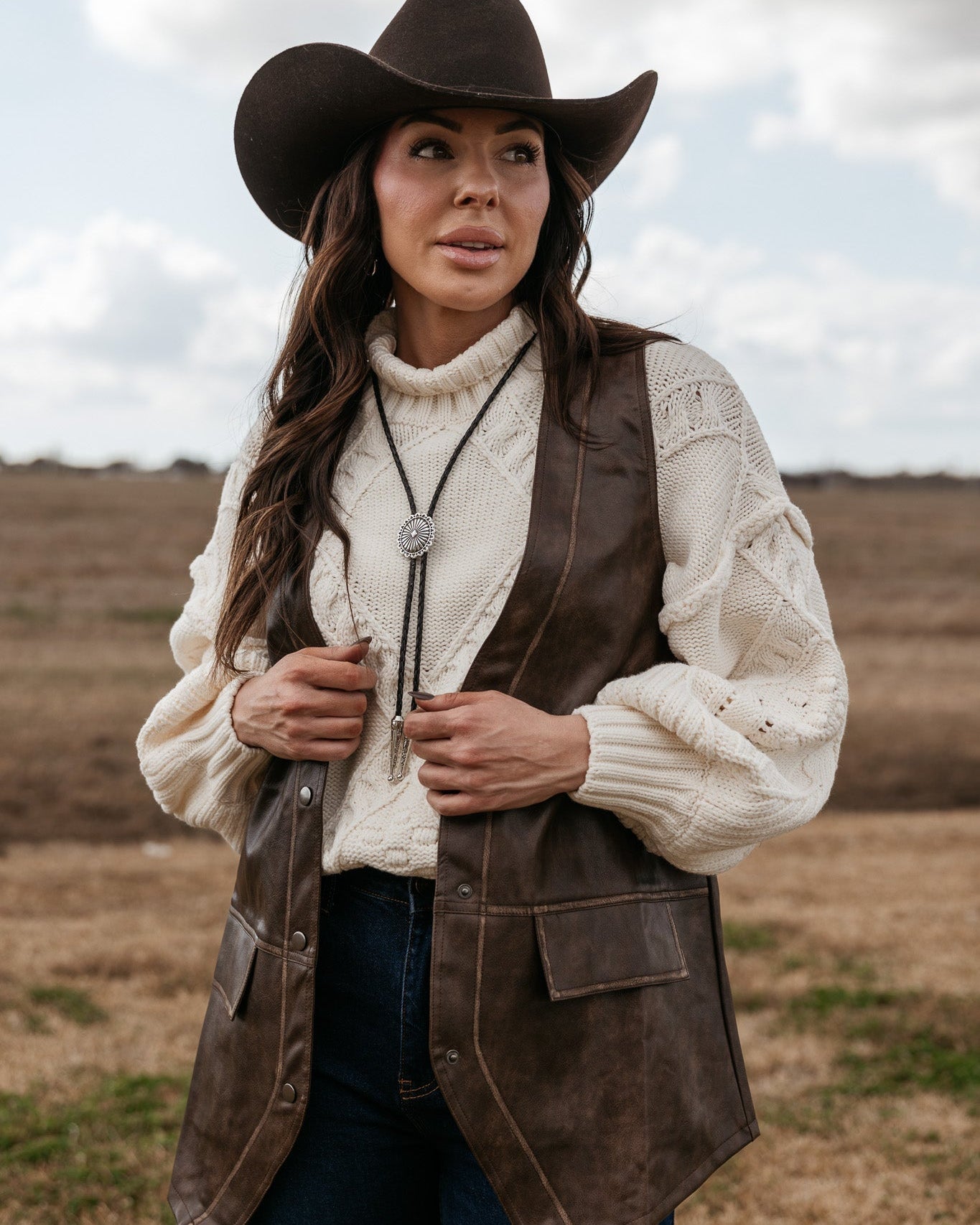 Woman in western hat, cream sweater, brown vest, and bolo tie standing outdoors