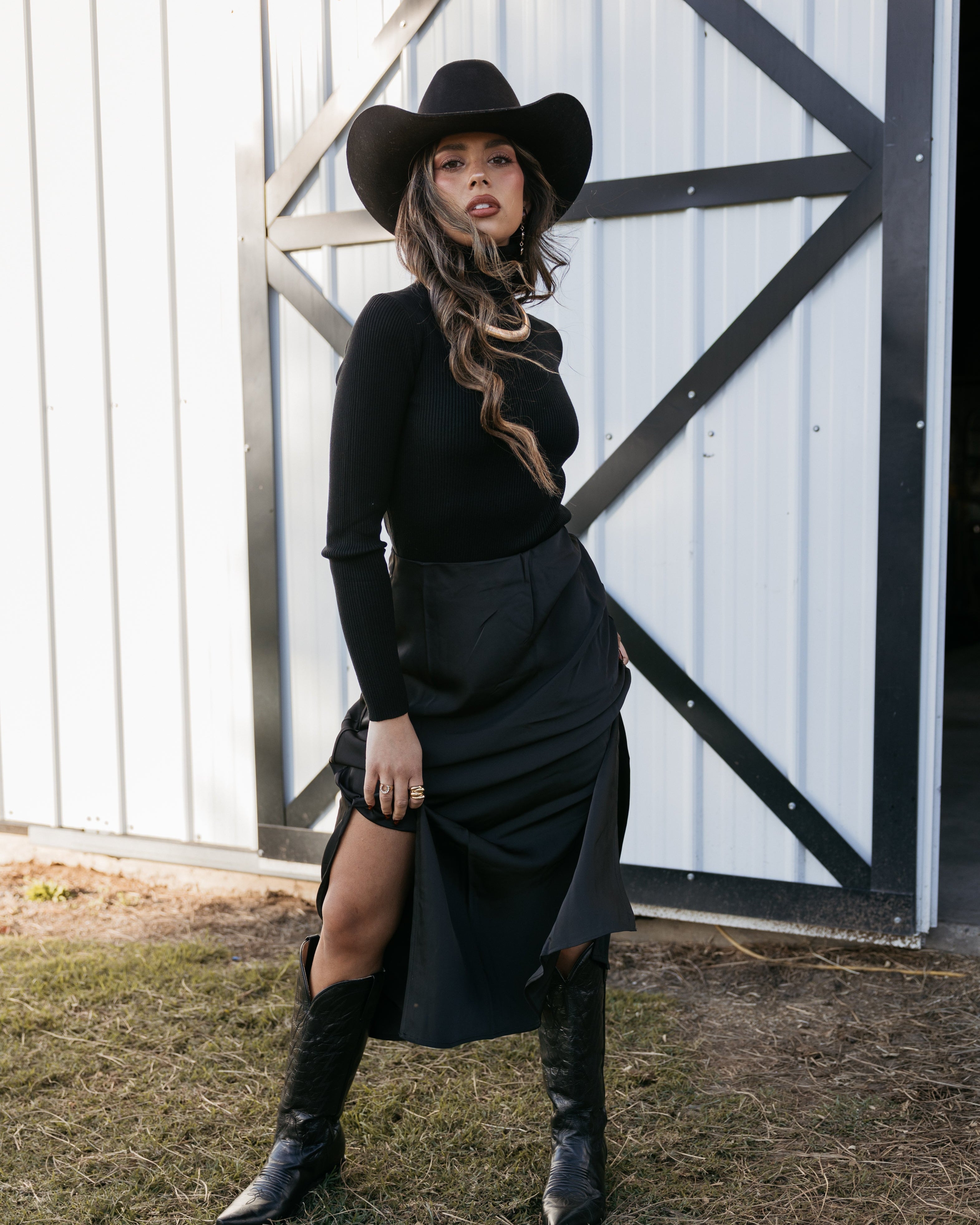 Woman in black cowboy hat, black long sleeve top, skirt, and boots posing in front of barn