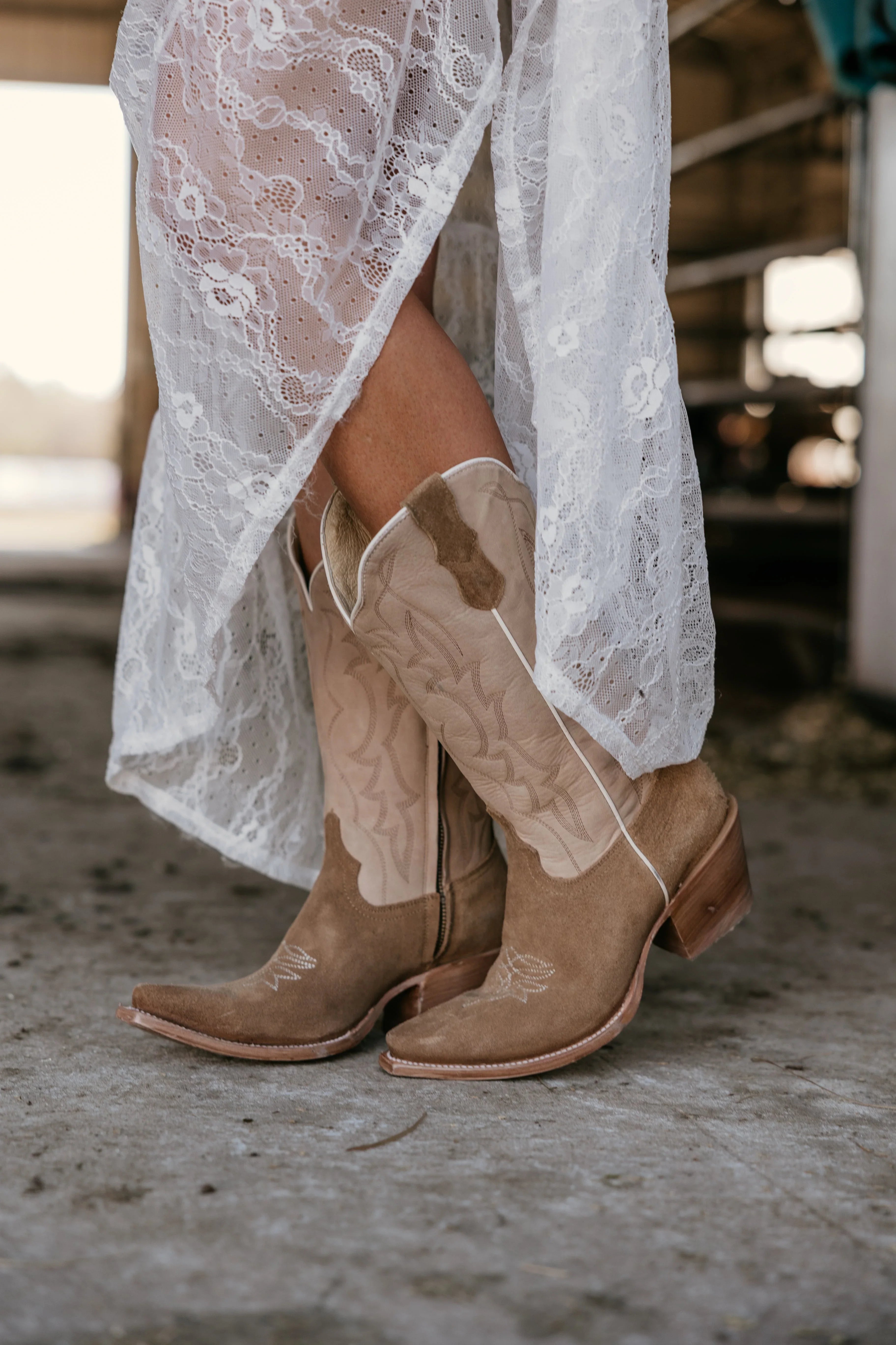 Tan cowboy boots worn with a white lace dress on a concrete floor.