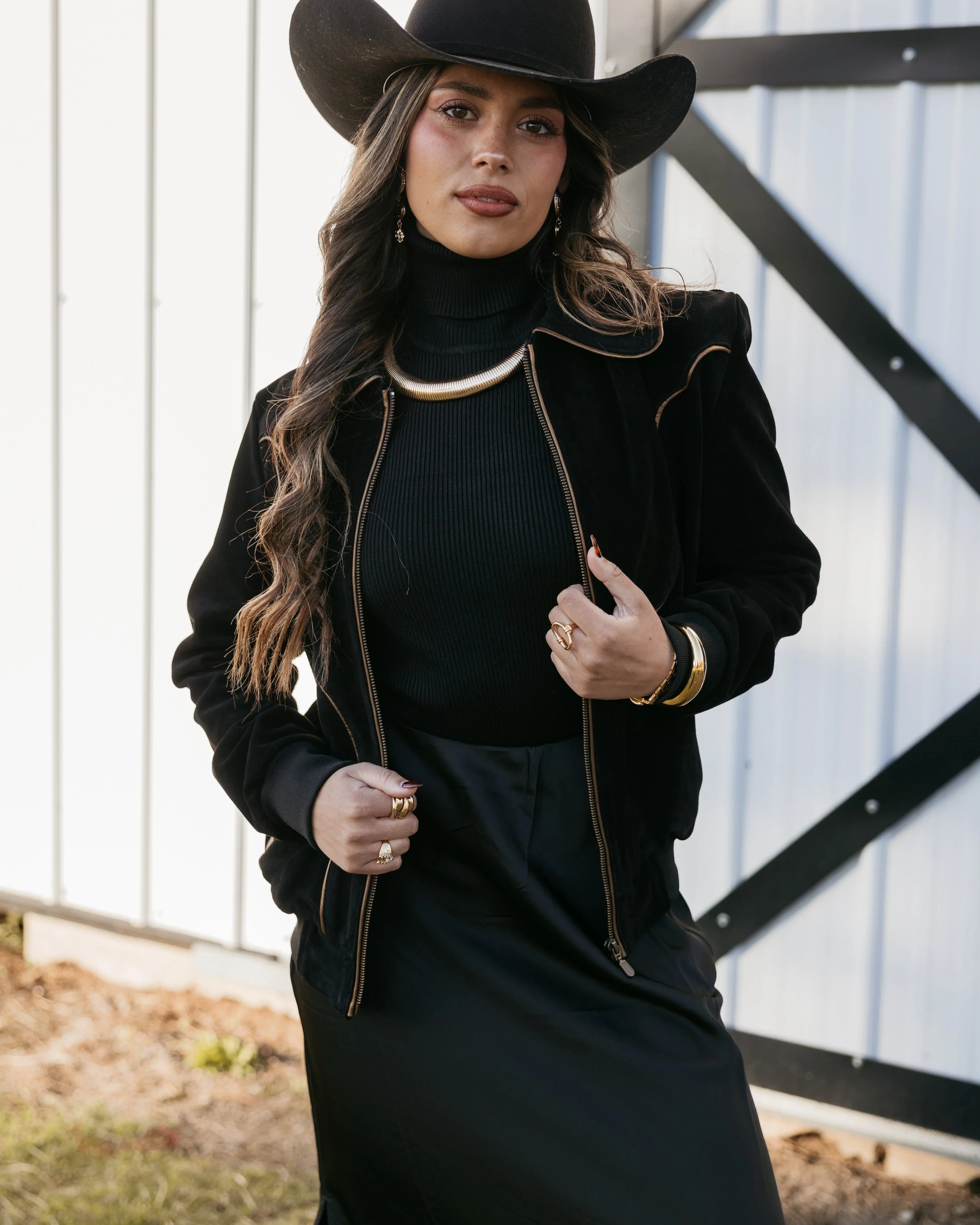 Woman in black cowgirl hat, western boho jacket, and jewelry posing by barn door