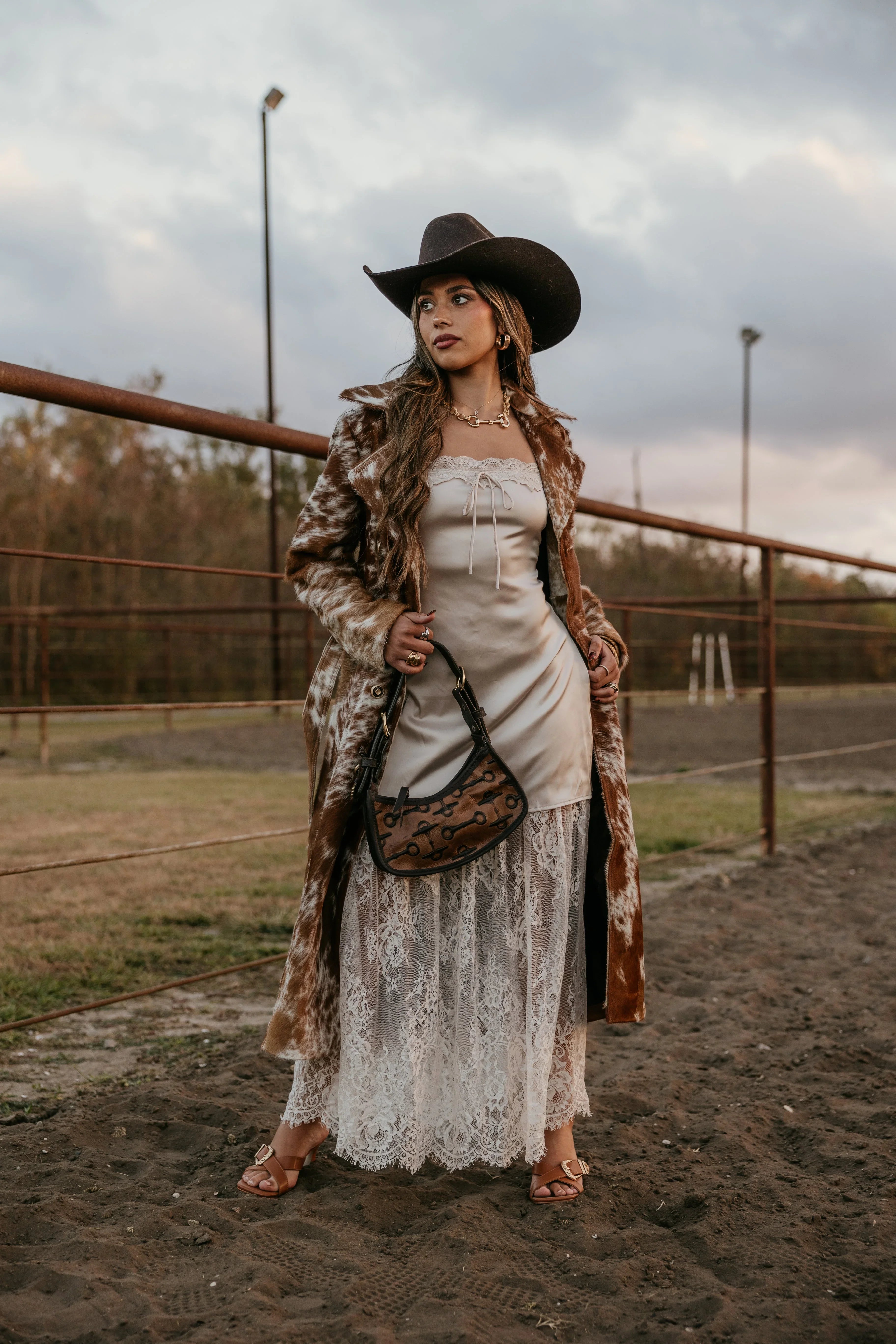 Woman in a white dress and brown coat standing in an outdoor setting with a cloudy sky.