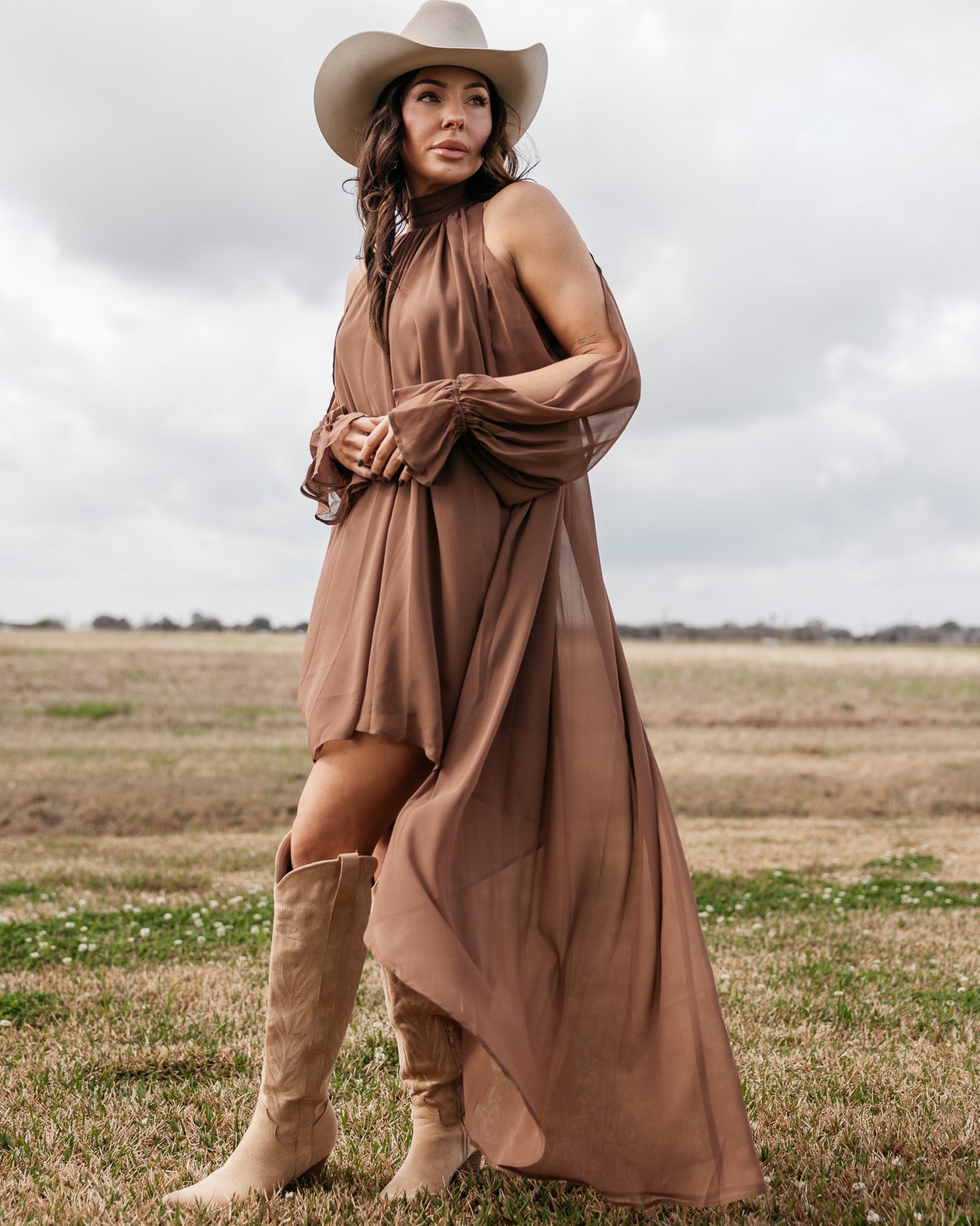 Woman in a brown western boho dress, cowgirl hat, and suede boots standing in a field.