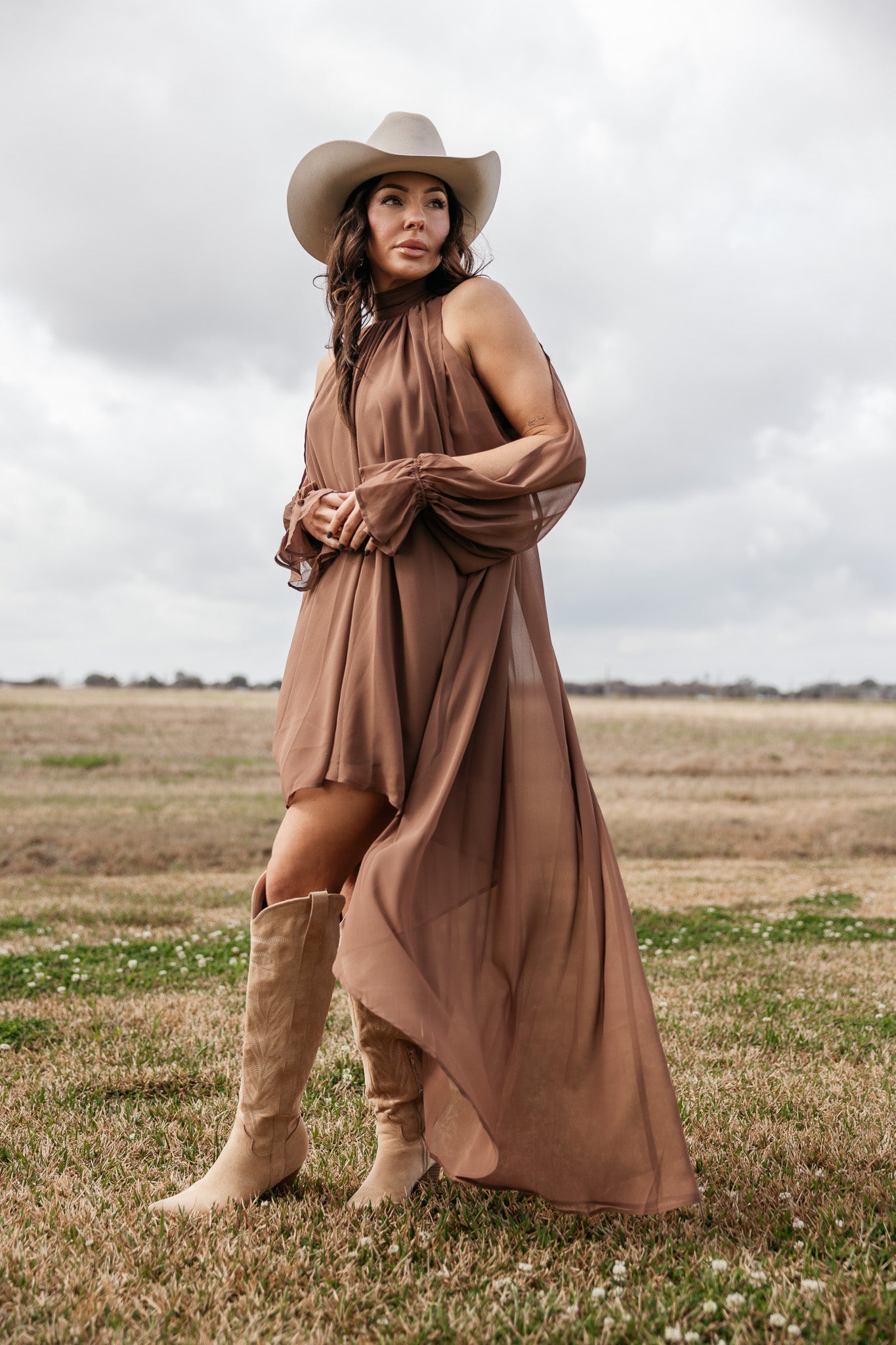 Woman in a brown western boho dress, cowgirl hat, and suede boots standing in a field.