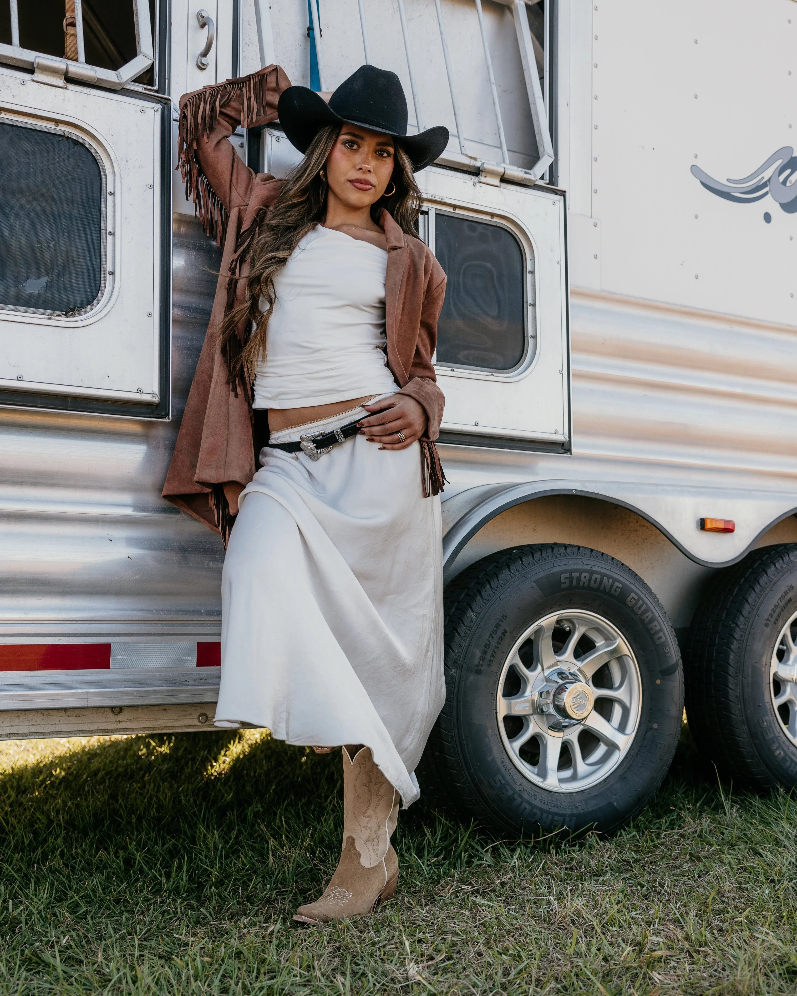 Woman in western boho outfit with suede fringe jacket, white skirt, and cowboy hat by horse trailer