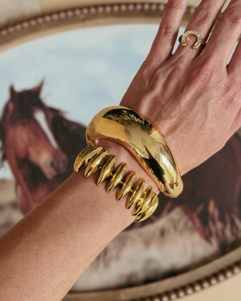 Gold western jewelry, including chunky bracelets and a horseshoe ring, worn on a hand with a horse painting in the background.