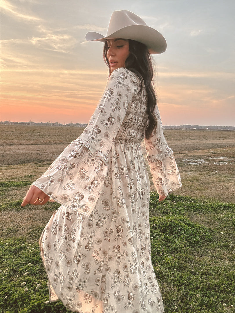 Woman in a floral western boho dress and cowboy hat standing in a field at sunset