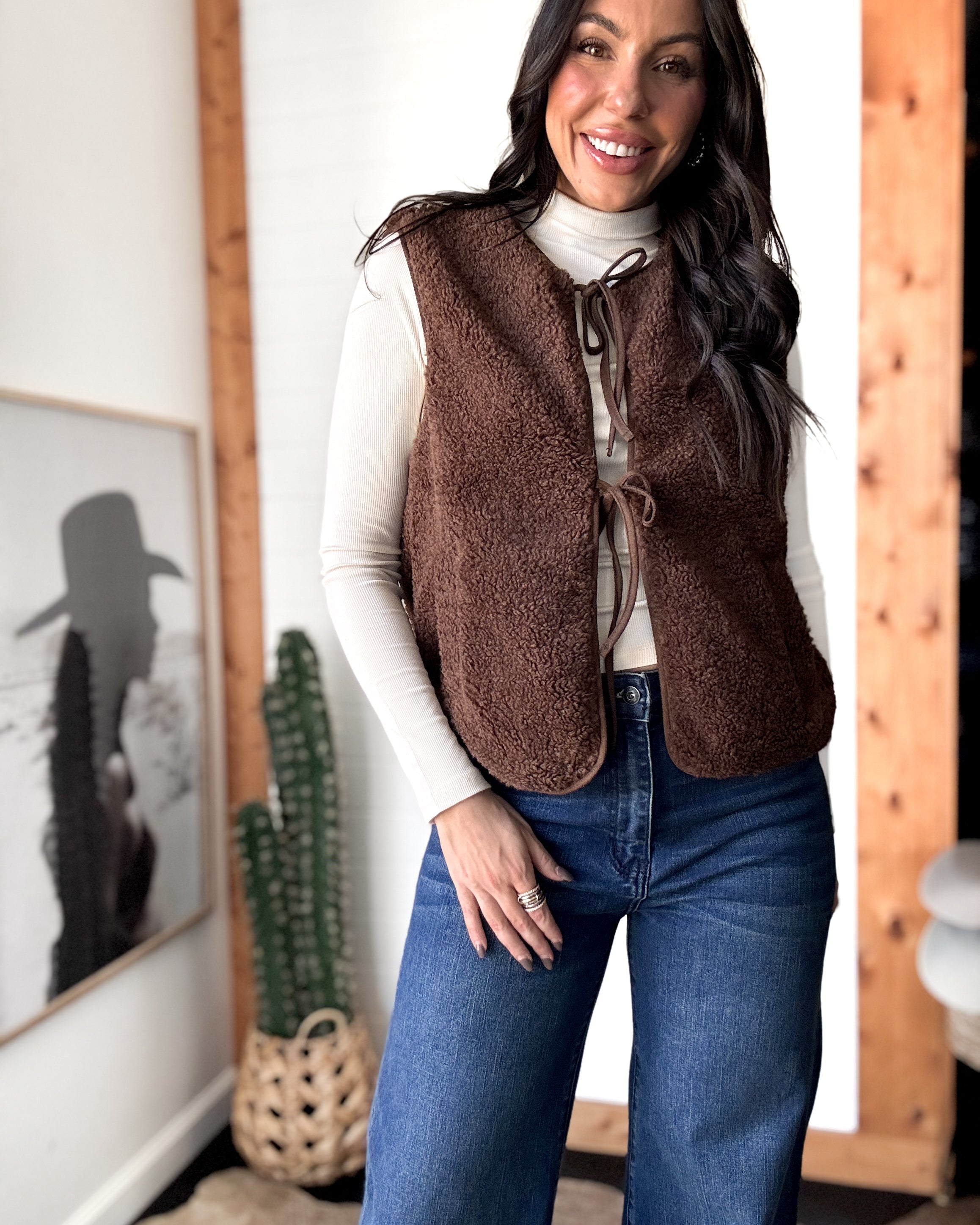 Woman wearing a brown vest over a white shirt and blue jeans indoors.